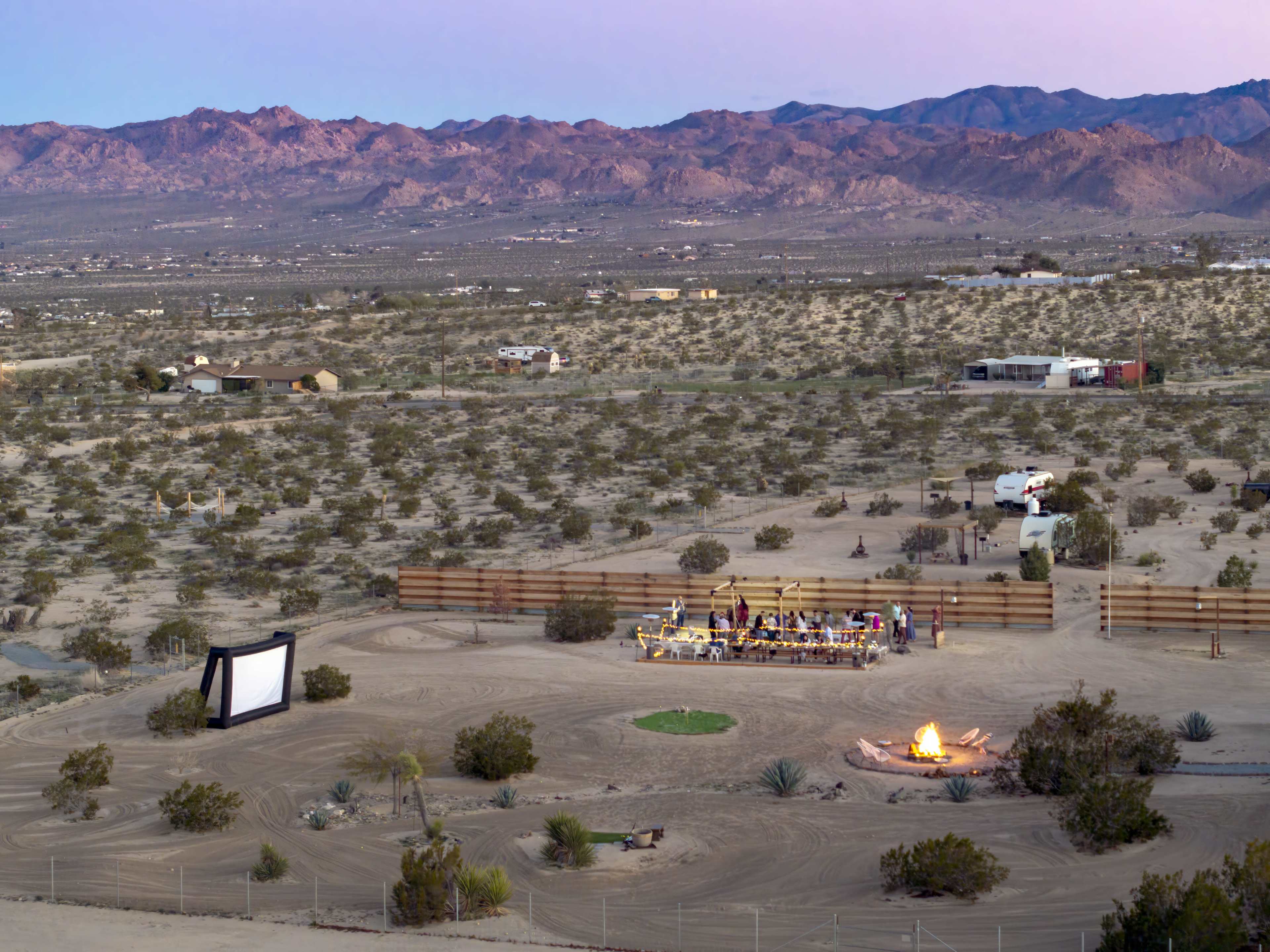 An outdoor movie screening is set up in a desert area, complete with a large screen, seating, and a campfire, surrounded by sparse vegetation and mountains in the background.