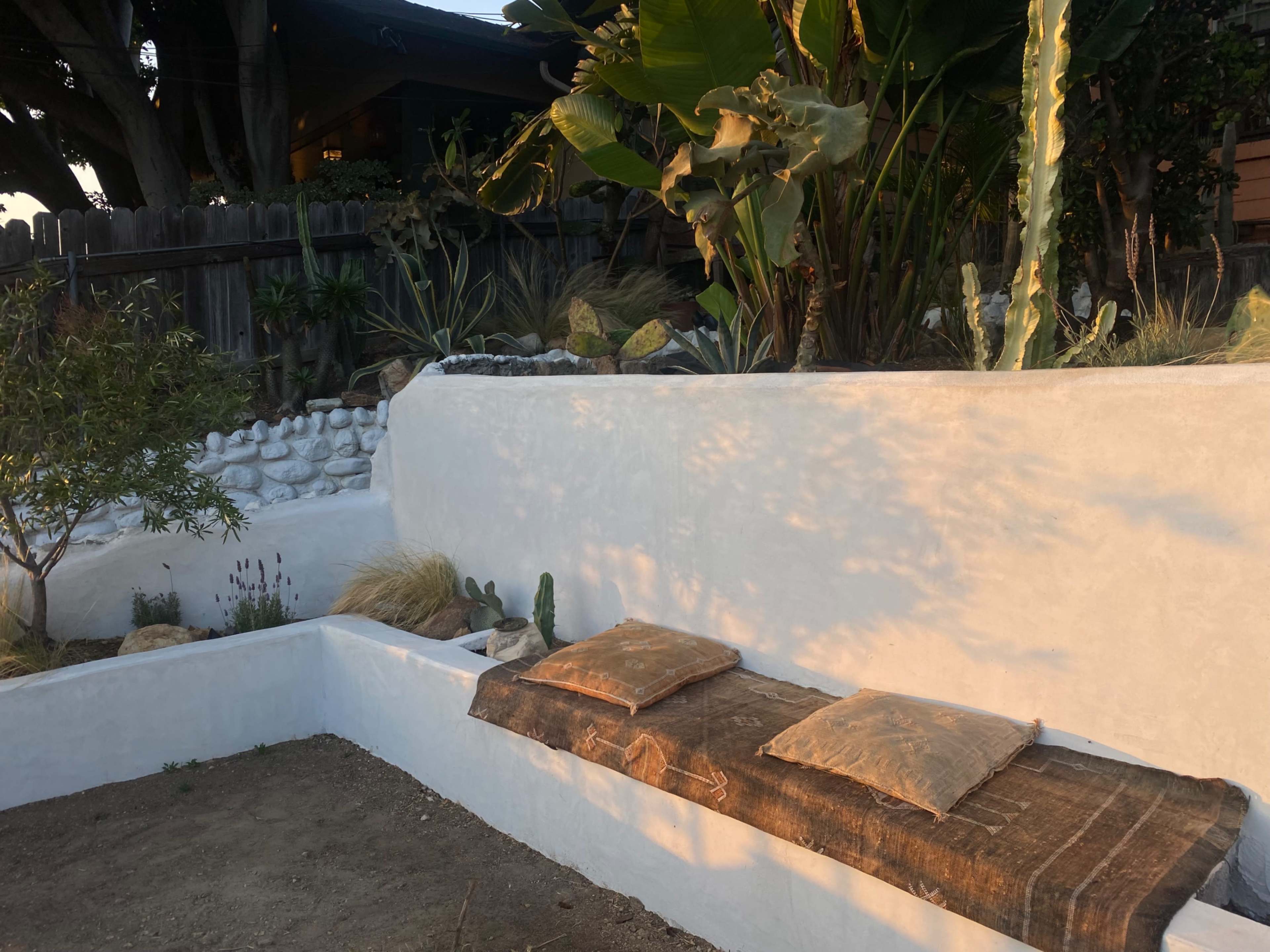A white stucco bench with two pillows sits against a garden wall adorned with various plants and a stone arrangement.