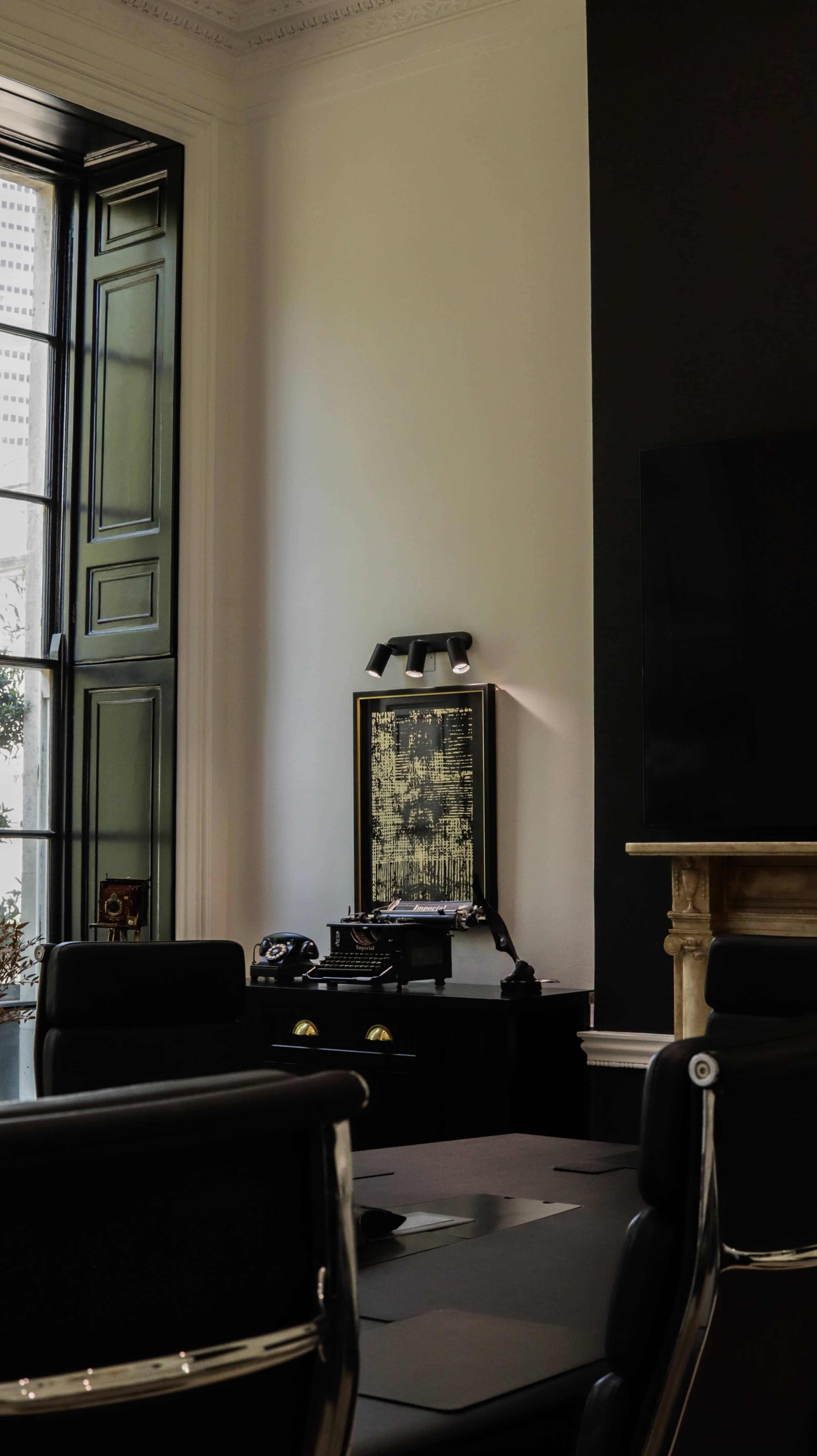 The image shows a stylish office space featuring a dark wall, a vintage typewriter on a black desk, and large windows with green shutters.