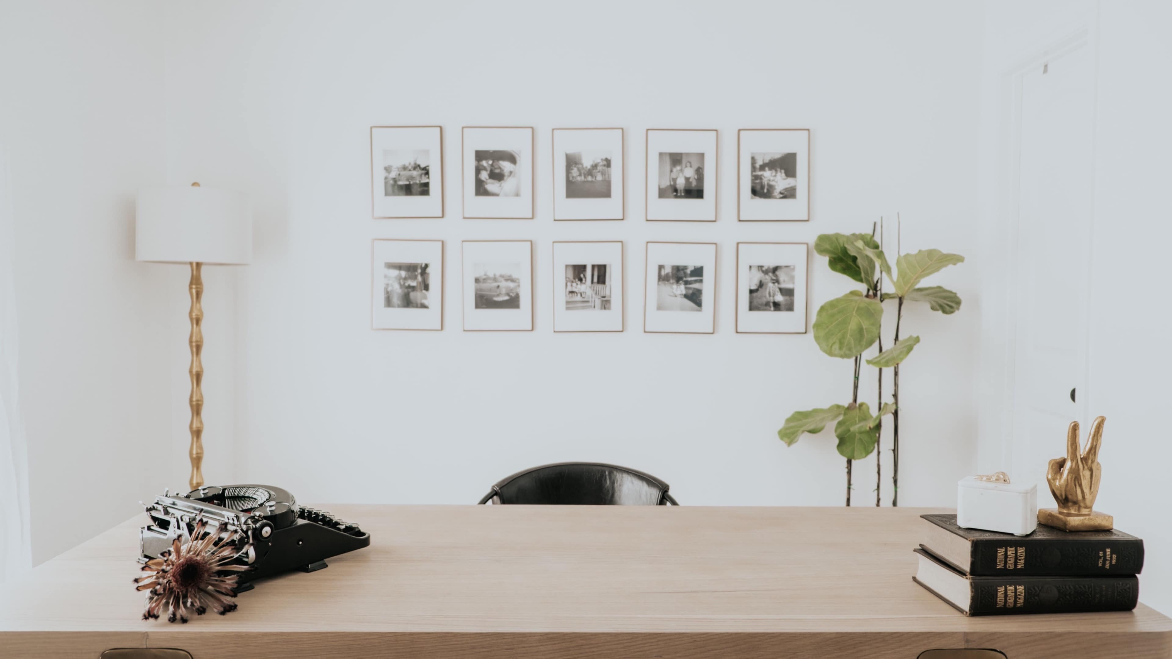 A minimalist office features a wooden desk with a typewriter, books, and a decorative plant, alongside a wall adorned with framed black-and-white photographs.