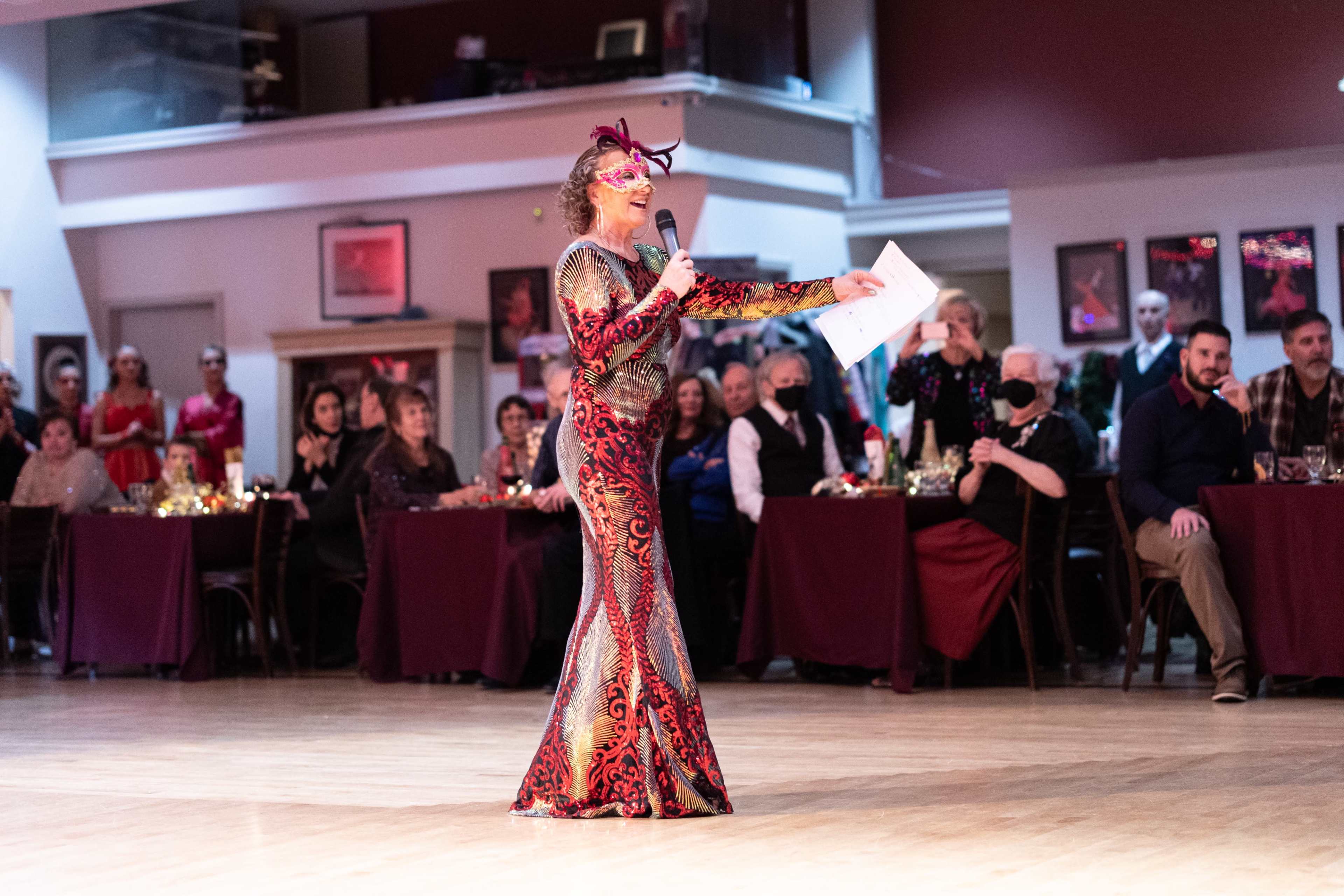 A performer in a brightly patterned gown holds a microphone while addressing an audience seated at tables in a banquet hall.