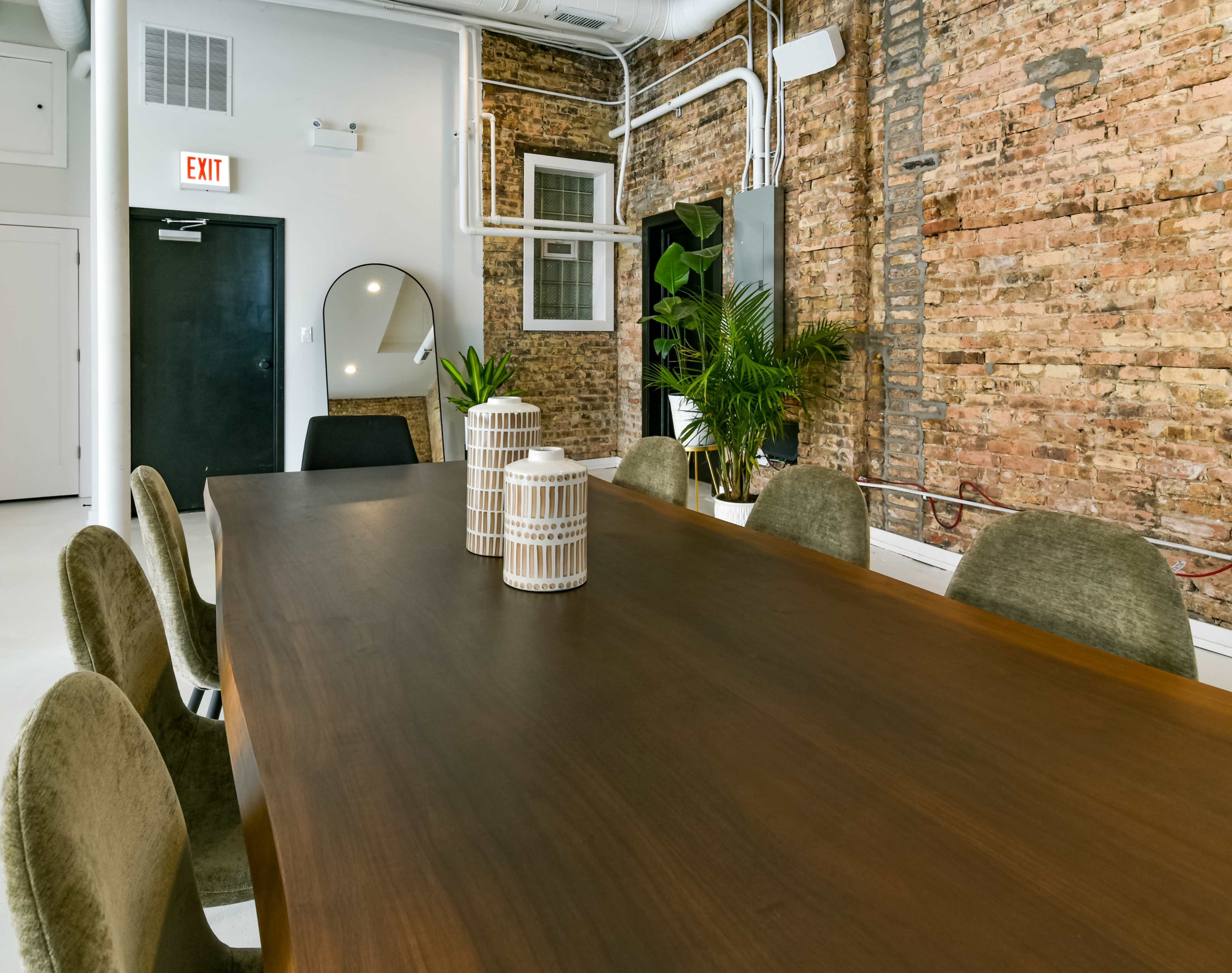 A long wooden dining table is set in a room with exposed brick walls and modern furnishings, featuring two decorative vases and a large mirror.