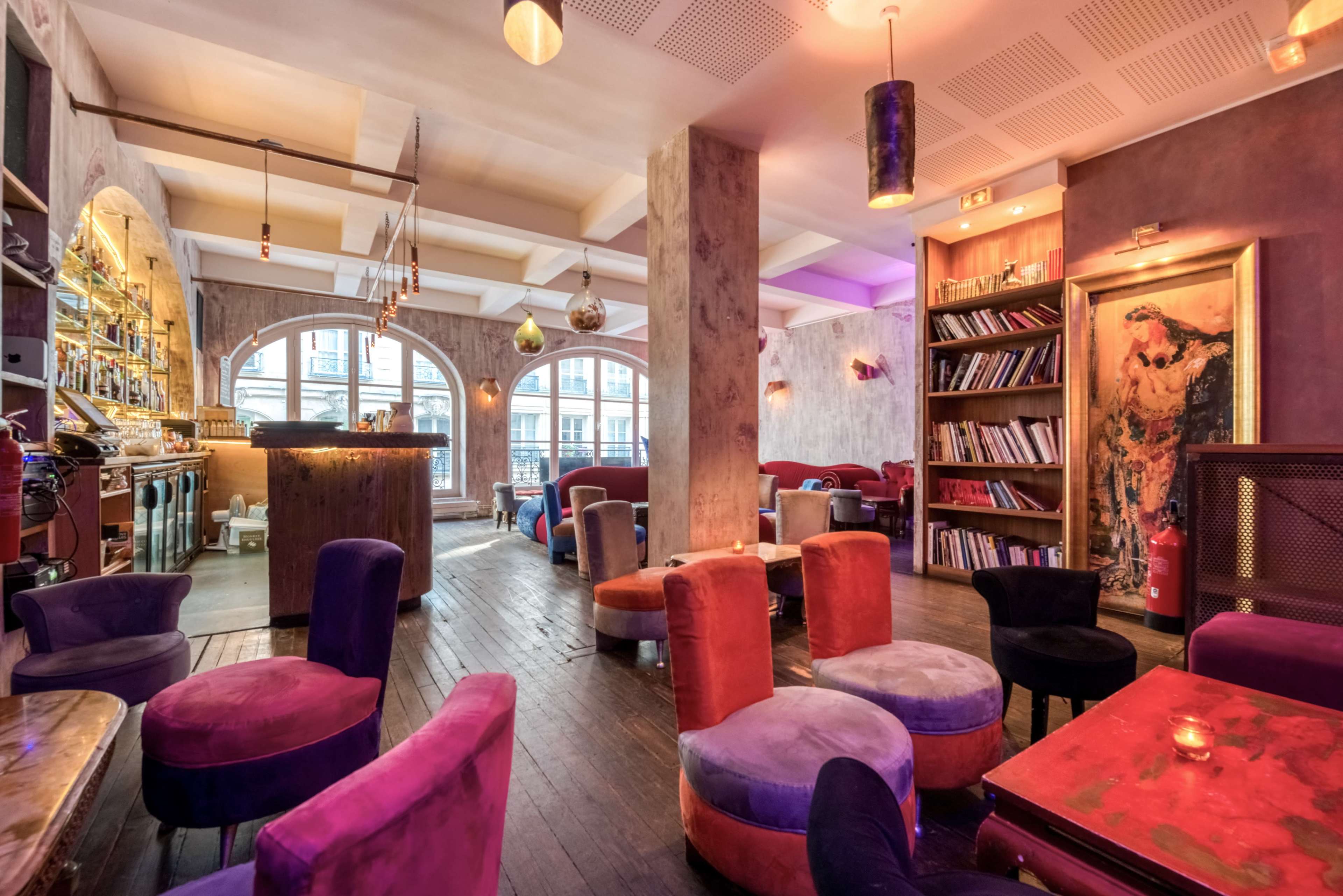 A cozy café interior with colorful armchairs, wooden floors, and arching windows that reveal a view of the outside.