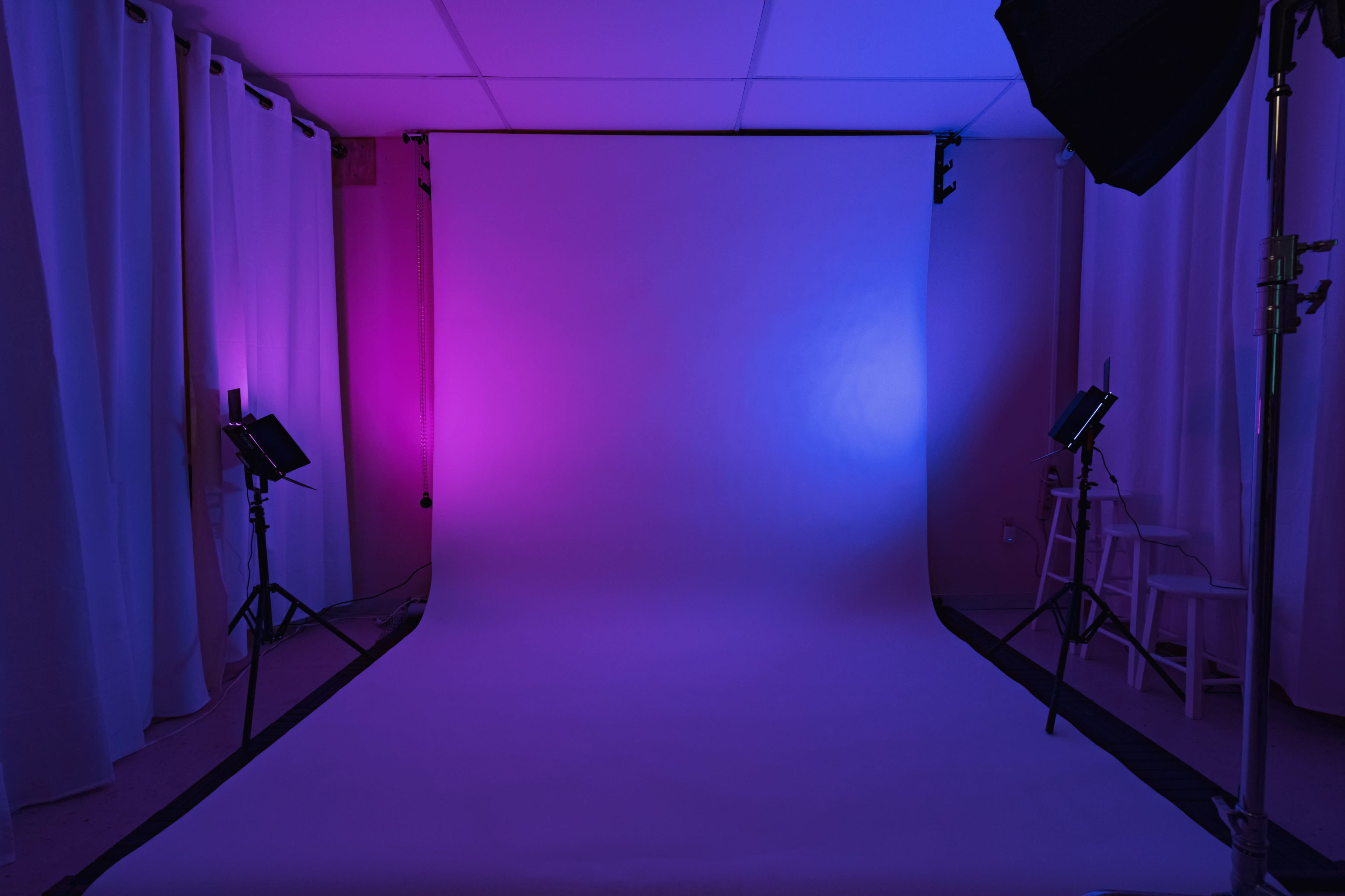 A photography studio with a backdrop illuminated by purple and blue lights, flanked by white curtains and lighting equipment.