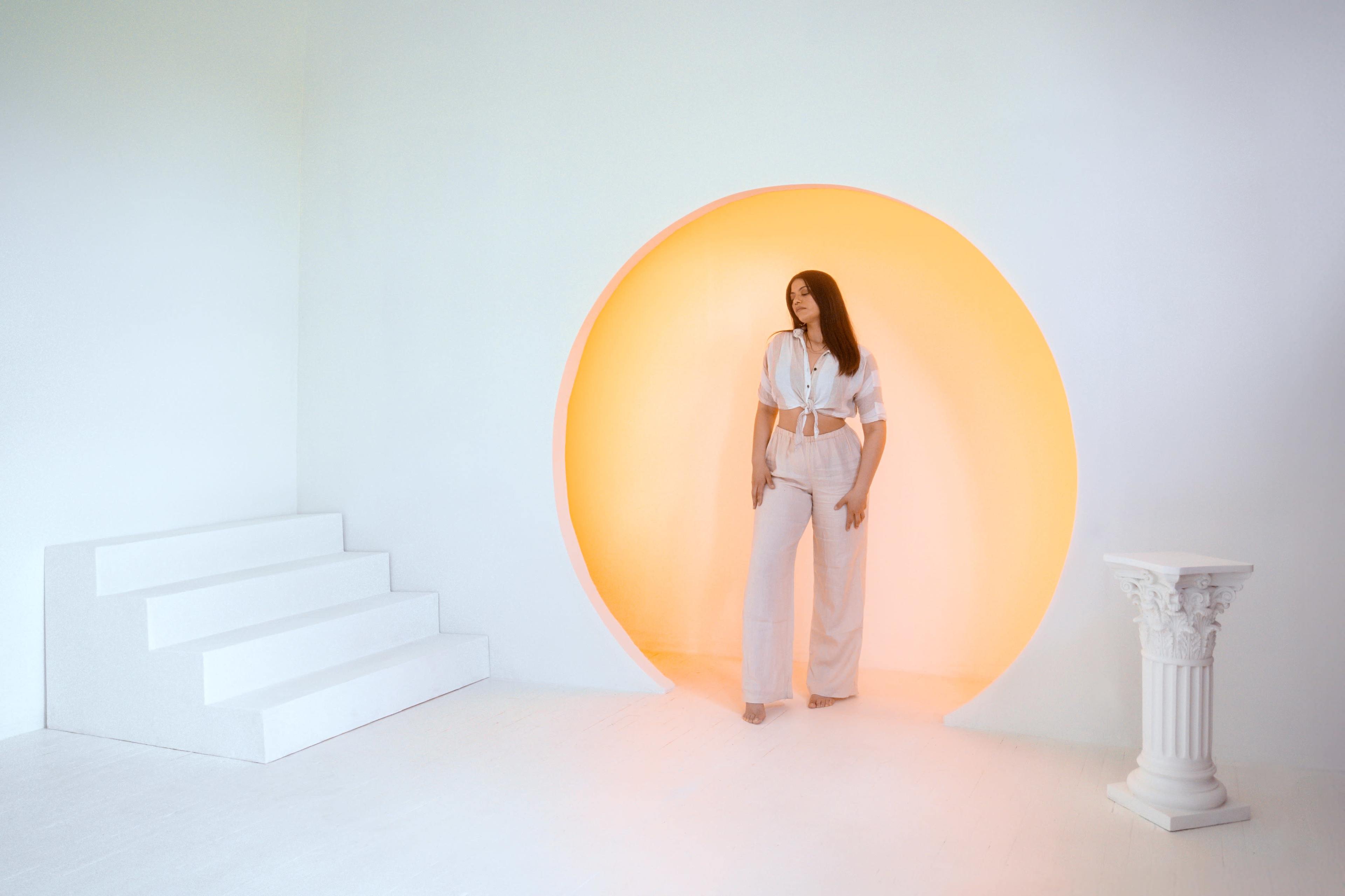 A woman in a white outfit stands inside a circular orange-lit alcove, with white steps leading up to it and a classical column beside her.