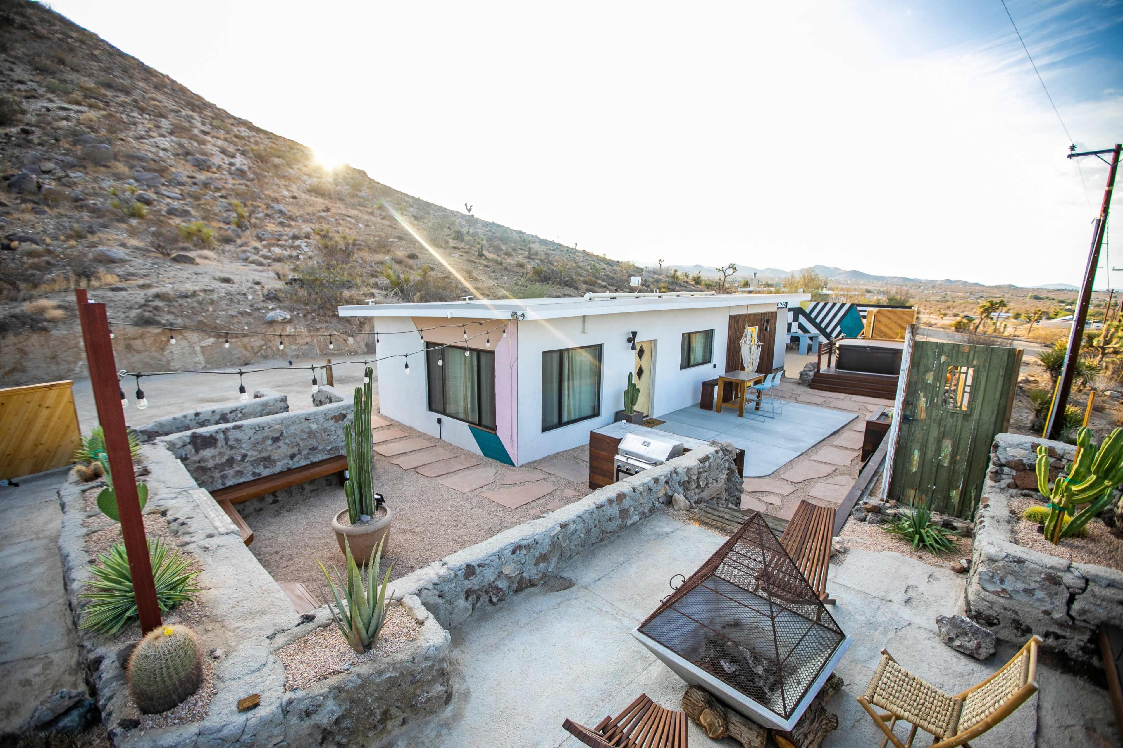 The image shows a modern house with a flat roof surrounded by desert terrain, featuring a patio area and various desert plants.