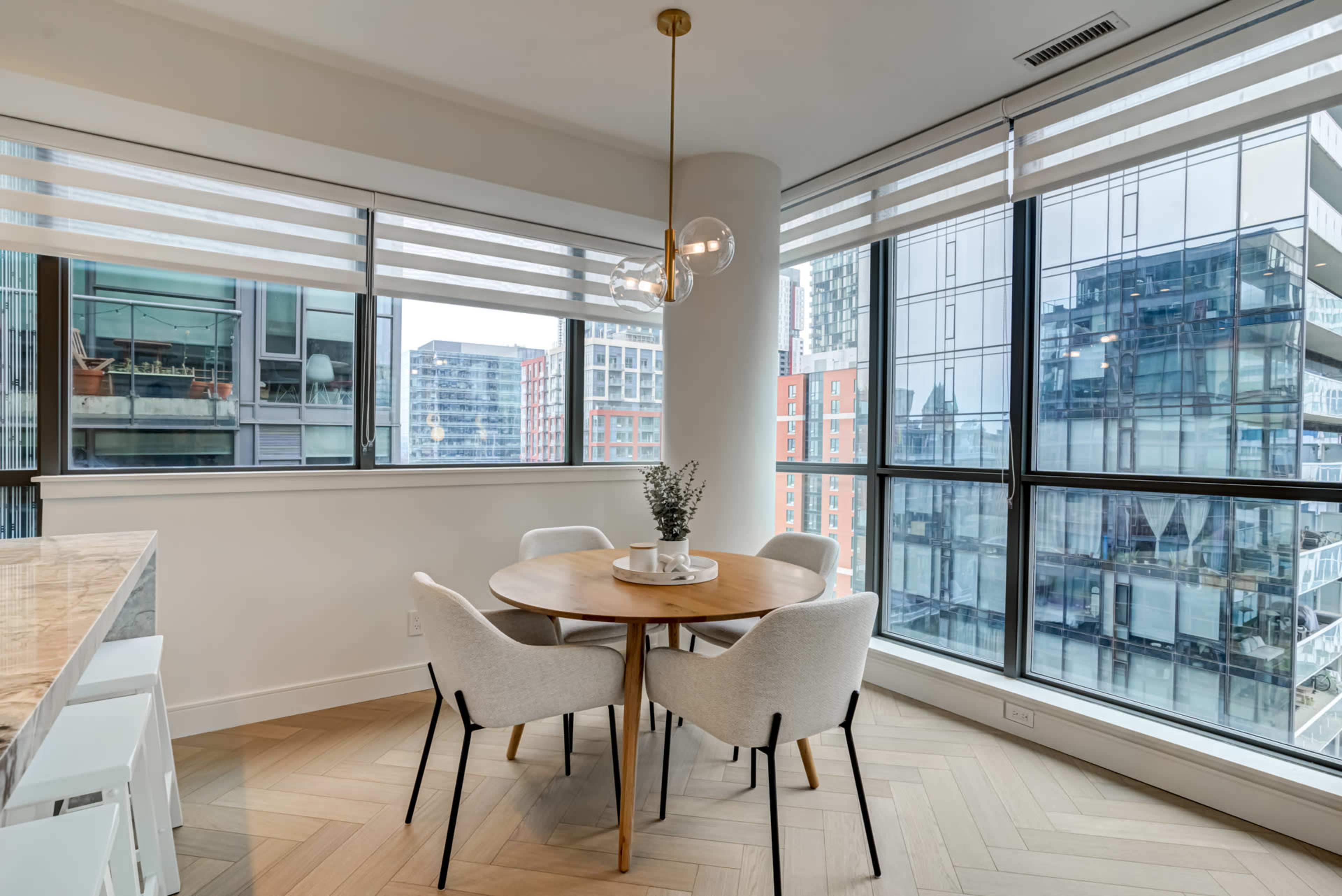 A round dining table with four chairs is positioned near a large window, showcasing a view of neighboring buildings.