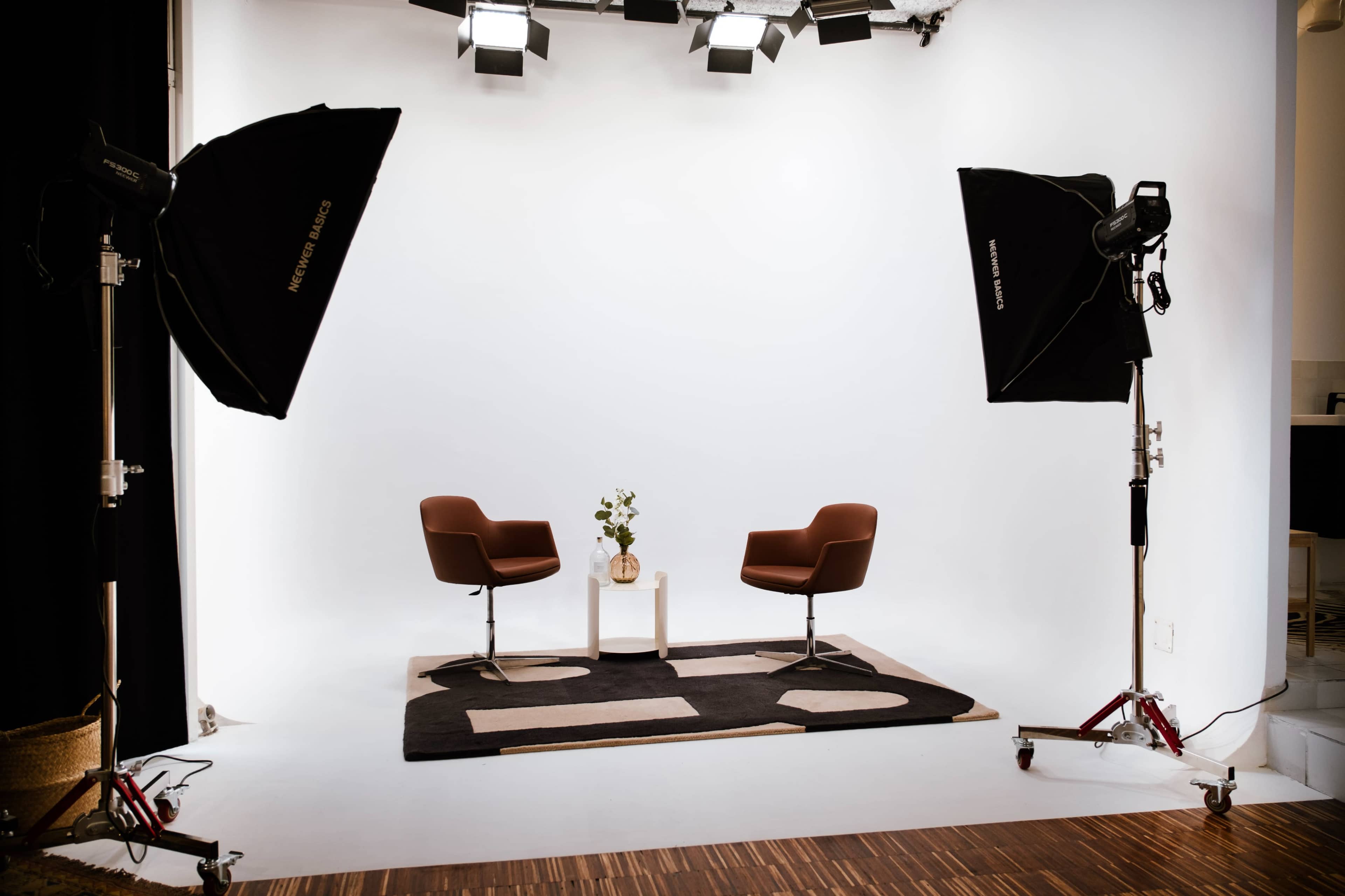 A studio setup features two brown chairs flanking a white table with a vase and a flower, illuminated by softbox lights against a white backdrop.