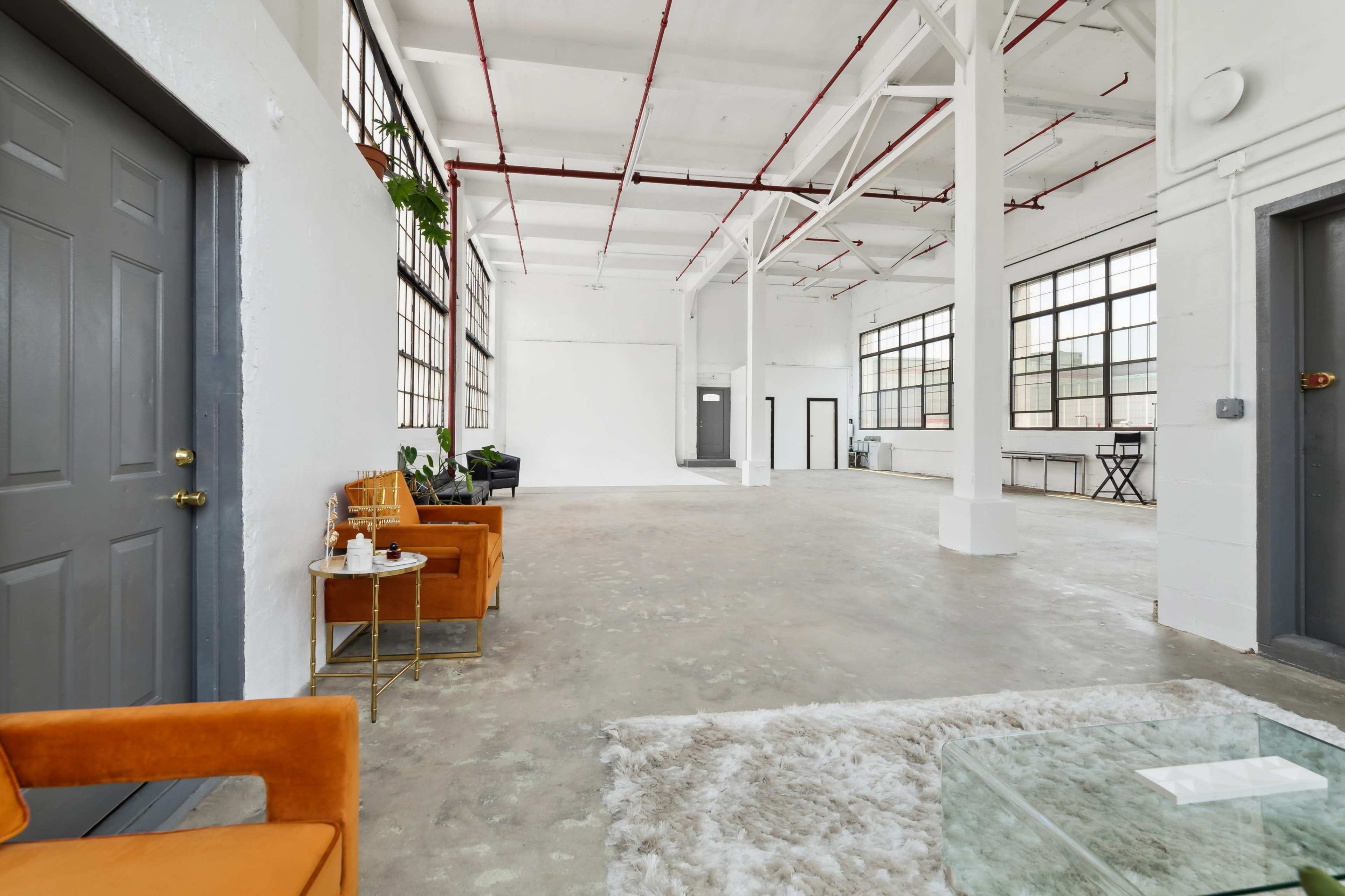 A spacious, well-lit industrial-style room with high ceilings, large windows, and minimal furniture.