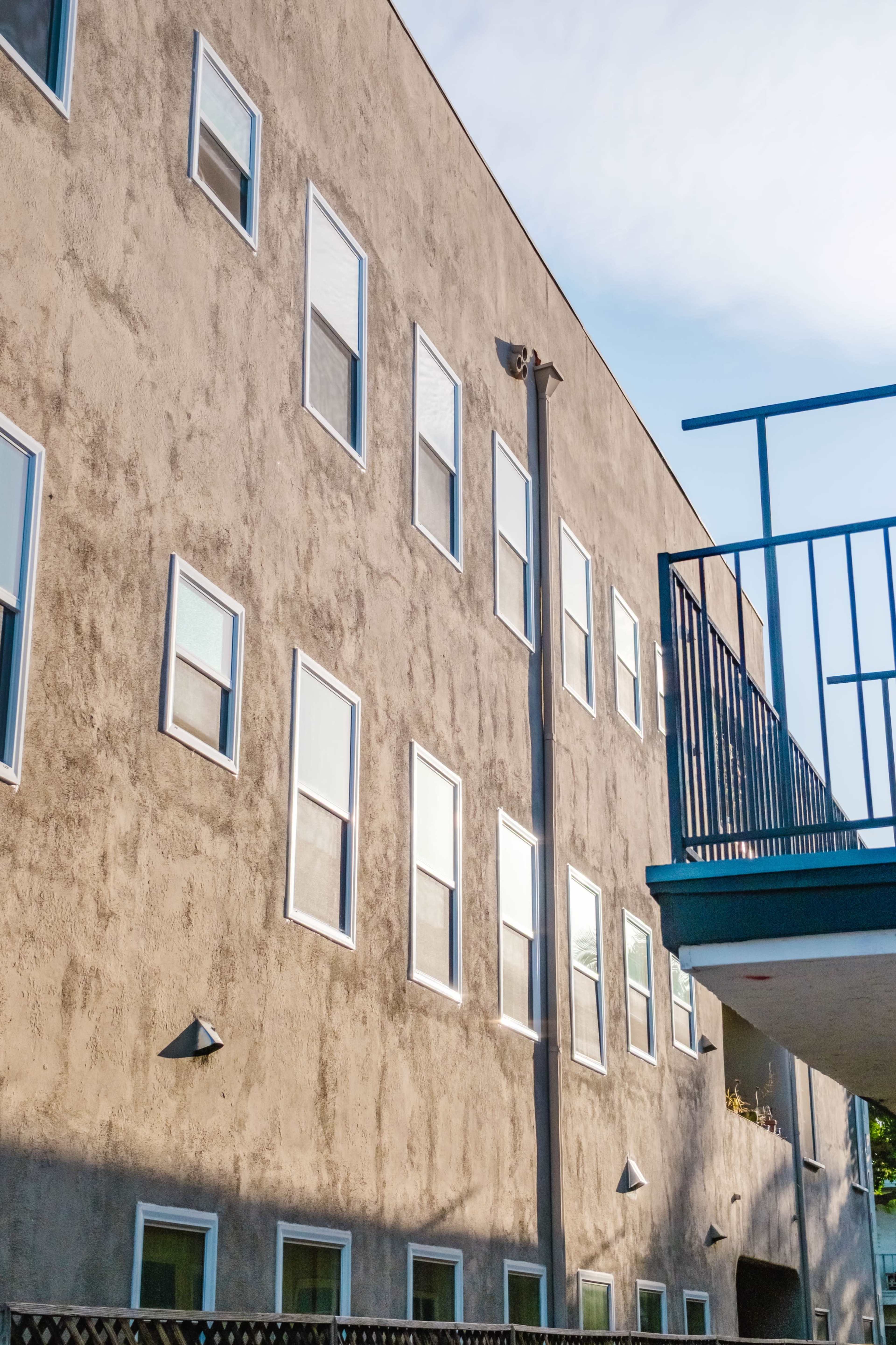 The image shows a multi-story building with a textured gray exterior and several rectangular windows, accompanied by a balcony on the right side.