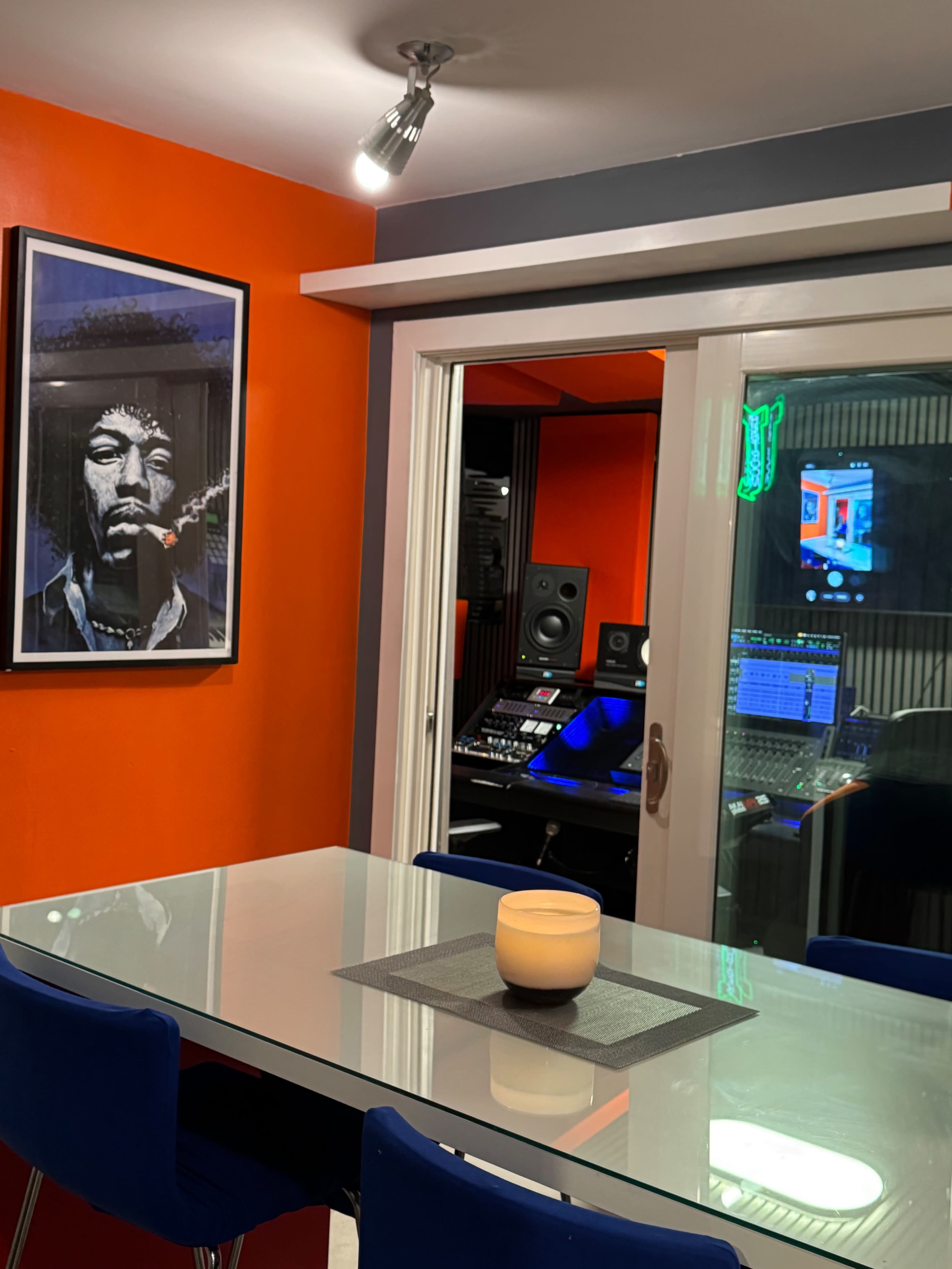 The image shows a modern recording studio visible through a glass door, featuring a glass table and a lit candle in the foreground.