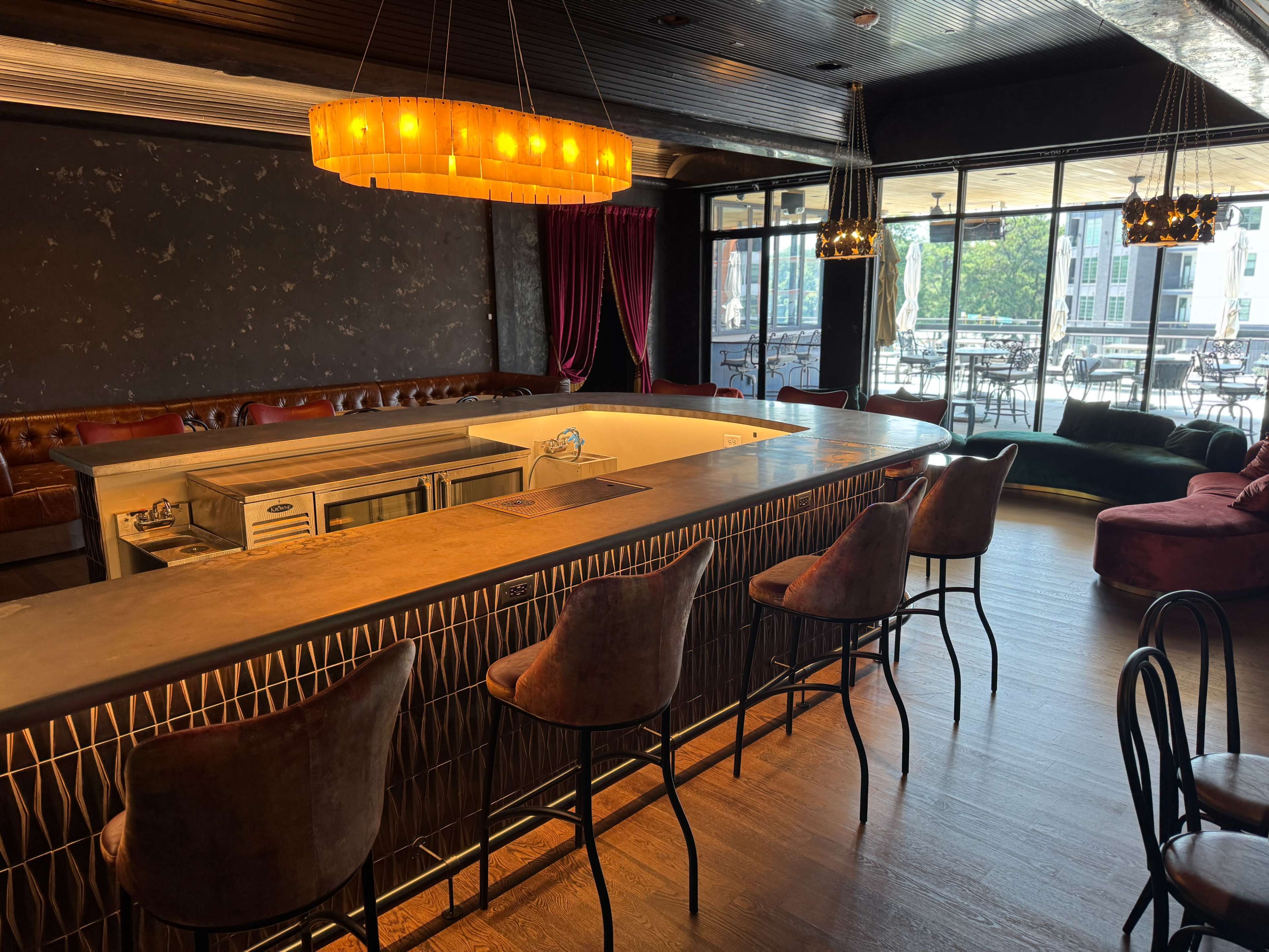 The image shows a stylish bar area with an illuminated chandelier, modern seating, and large windows overlooking an outdoor terrace.
