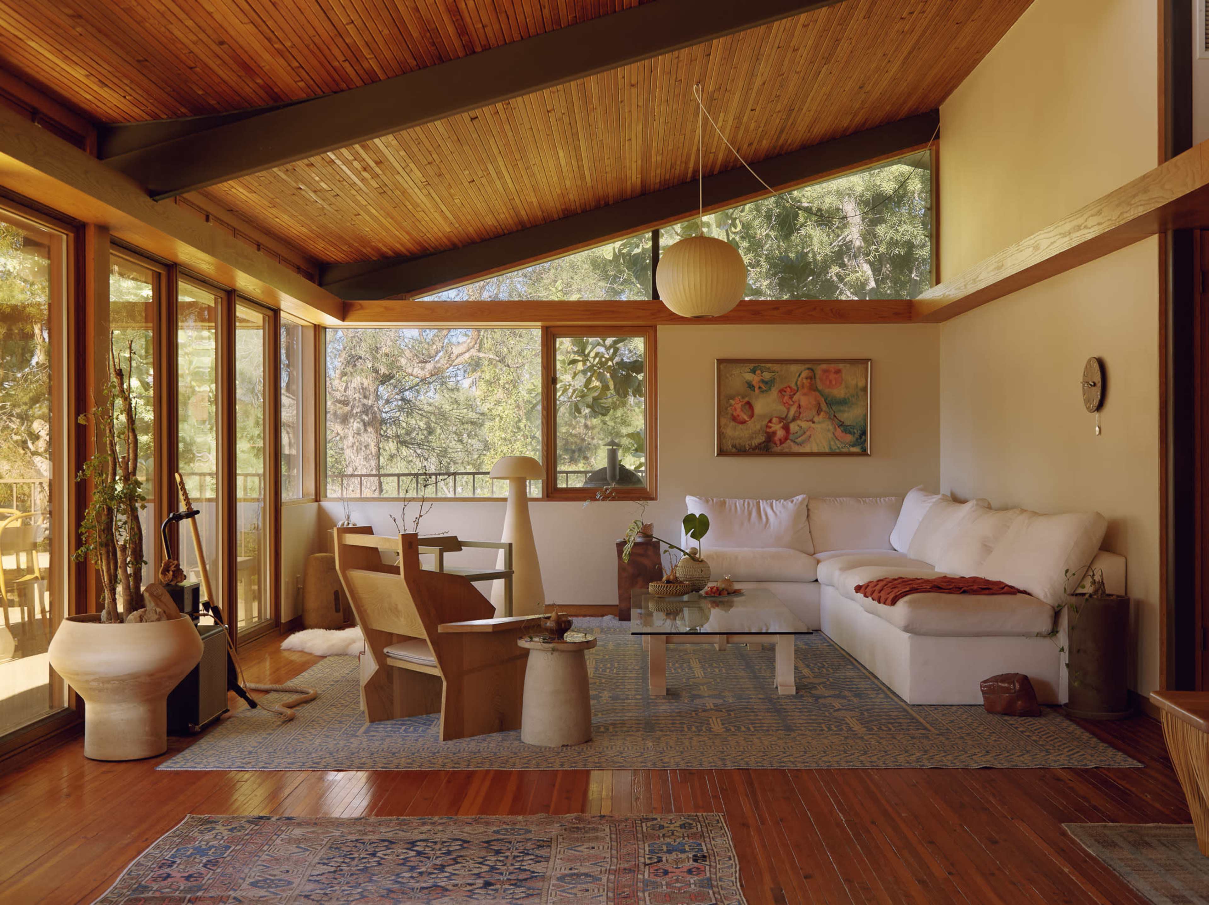 The scene features a spacious living room with large windows, wooden beams, and a mix of modern and rustic furniture, including a sectional sofa and a coffee table.