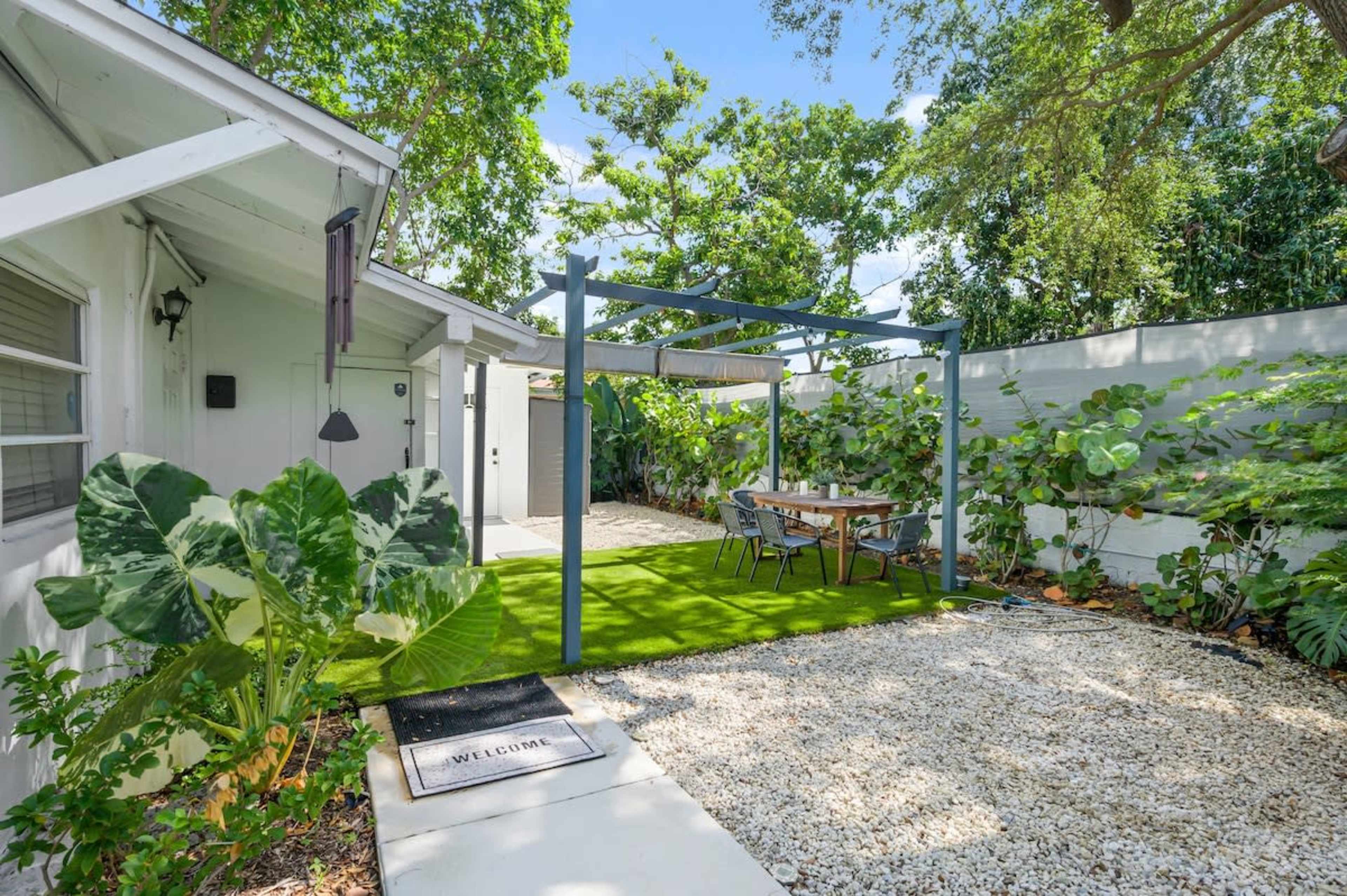 A small outdoor seating area with a table under a pergola, surrounded by greenery and gravel pathways next to a house.