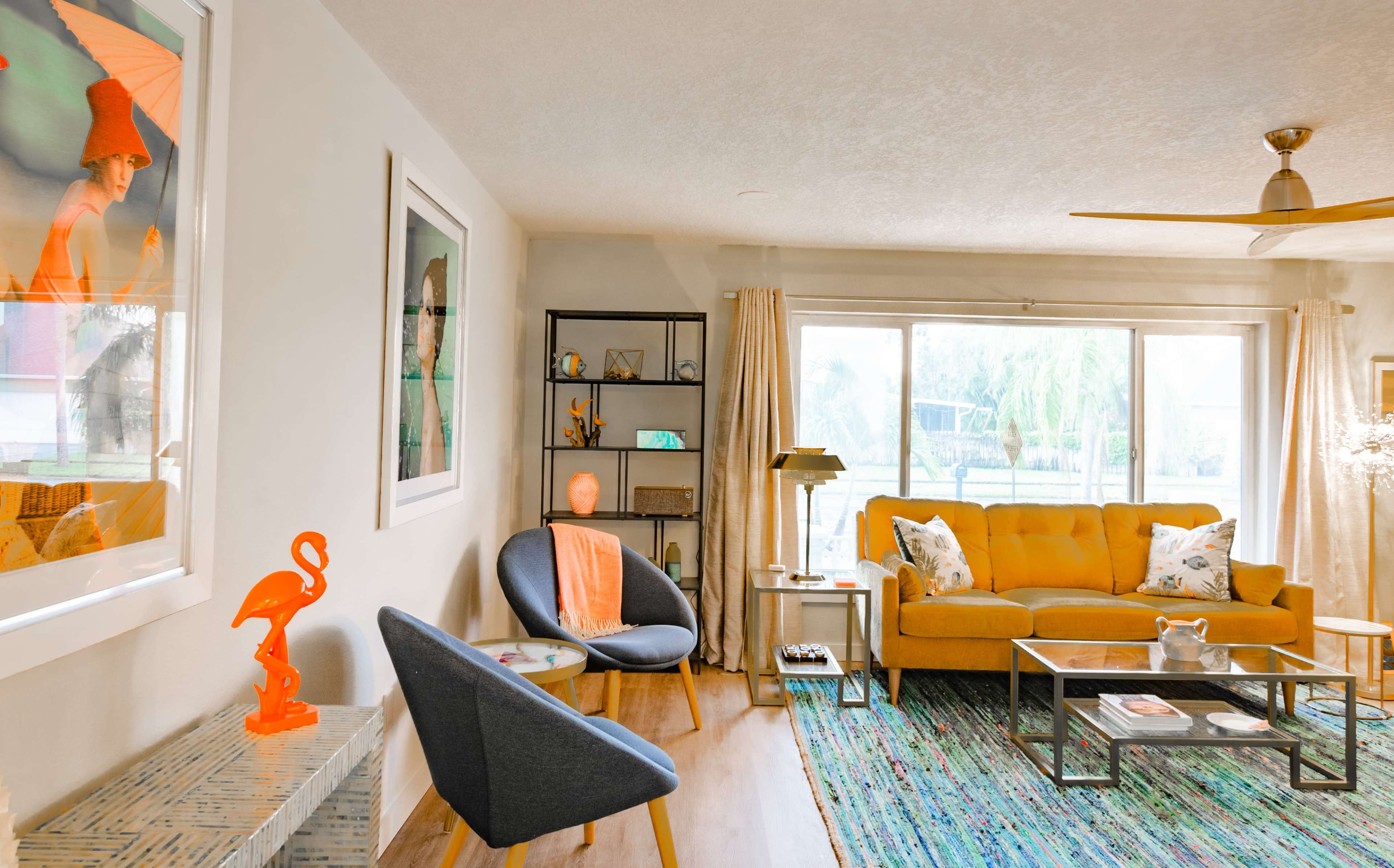 A modern living room featuring a yellow sofa, a blue armchair, large windows with soft curtains, and colorful artwork on the walls.
