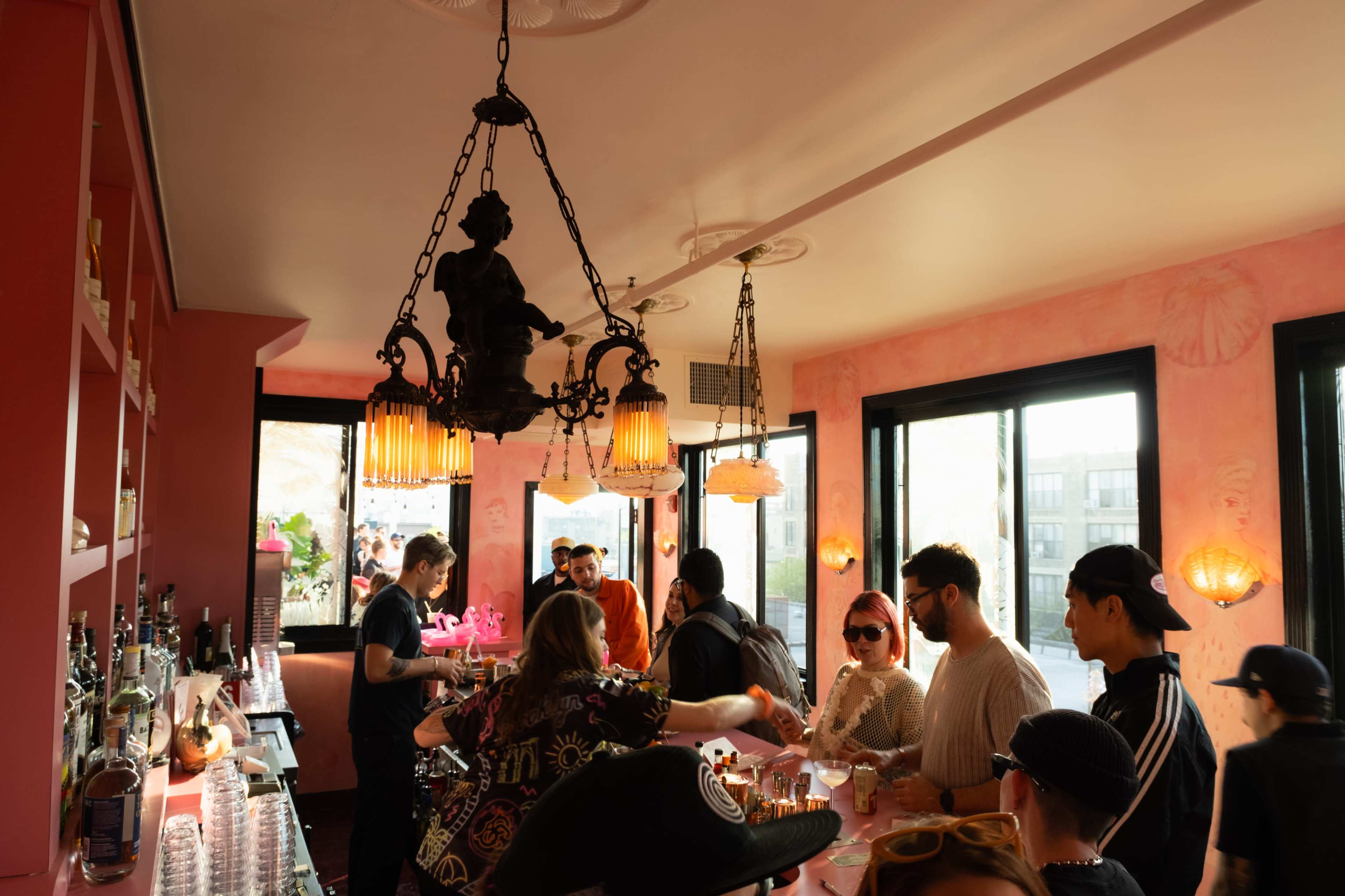 A group of people socialize at a brightly lit bar with pink walls and vintage chandeliers.