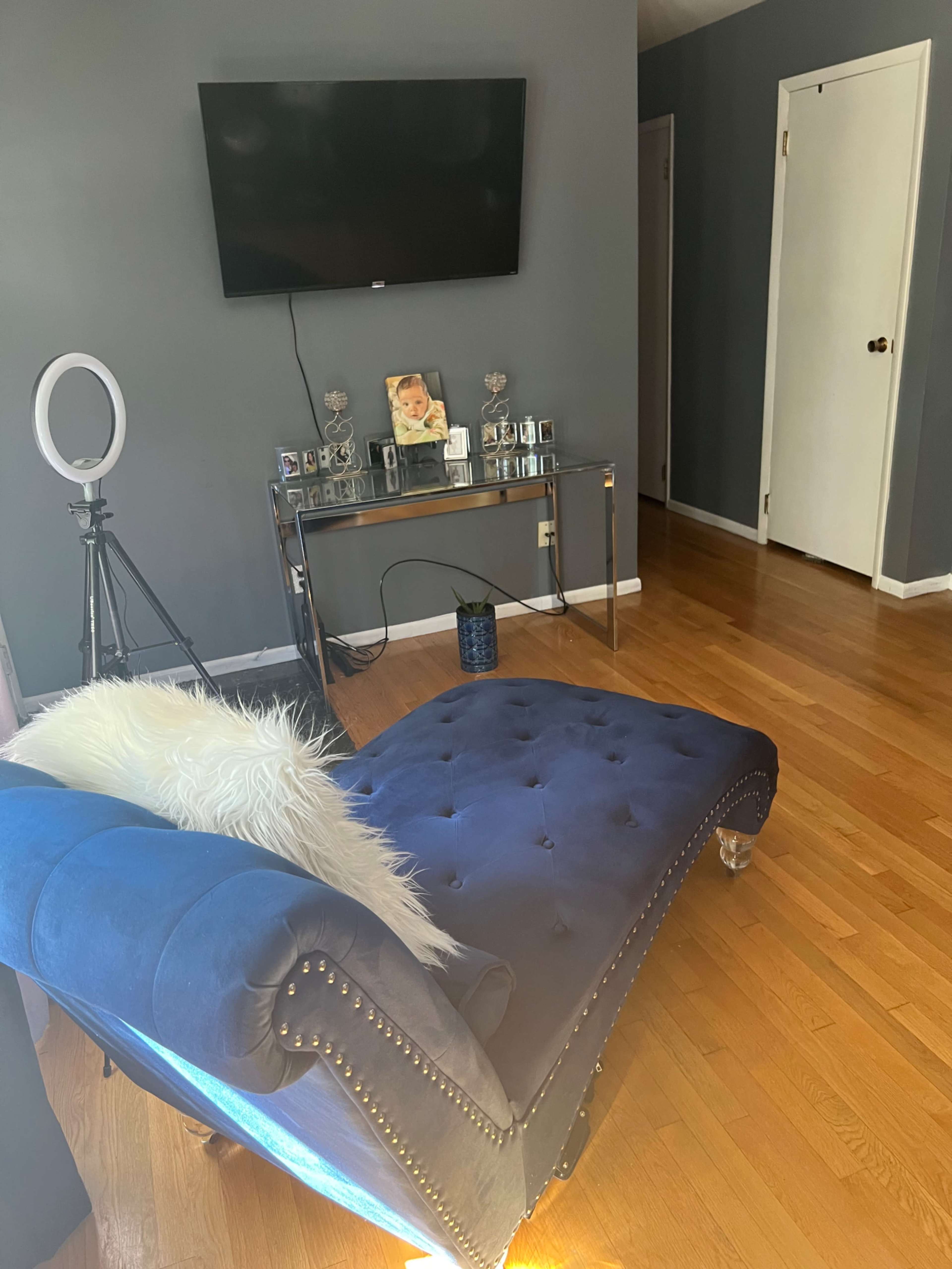 A blue chaise lounge is positioned in a well-lit room featuring a television on the wall and a glass side table displaying framed photos.