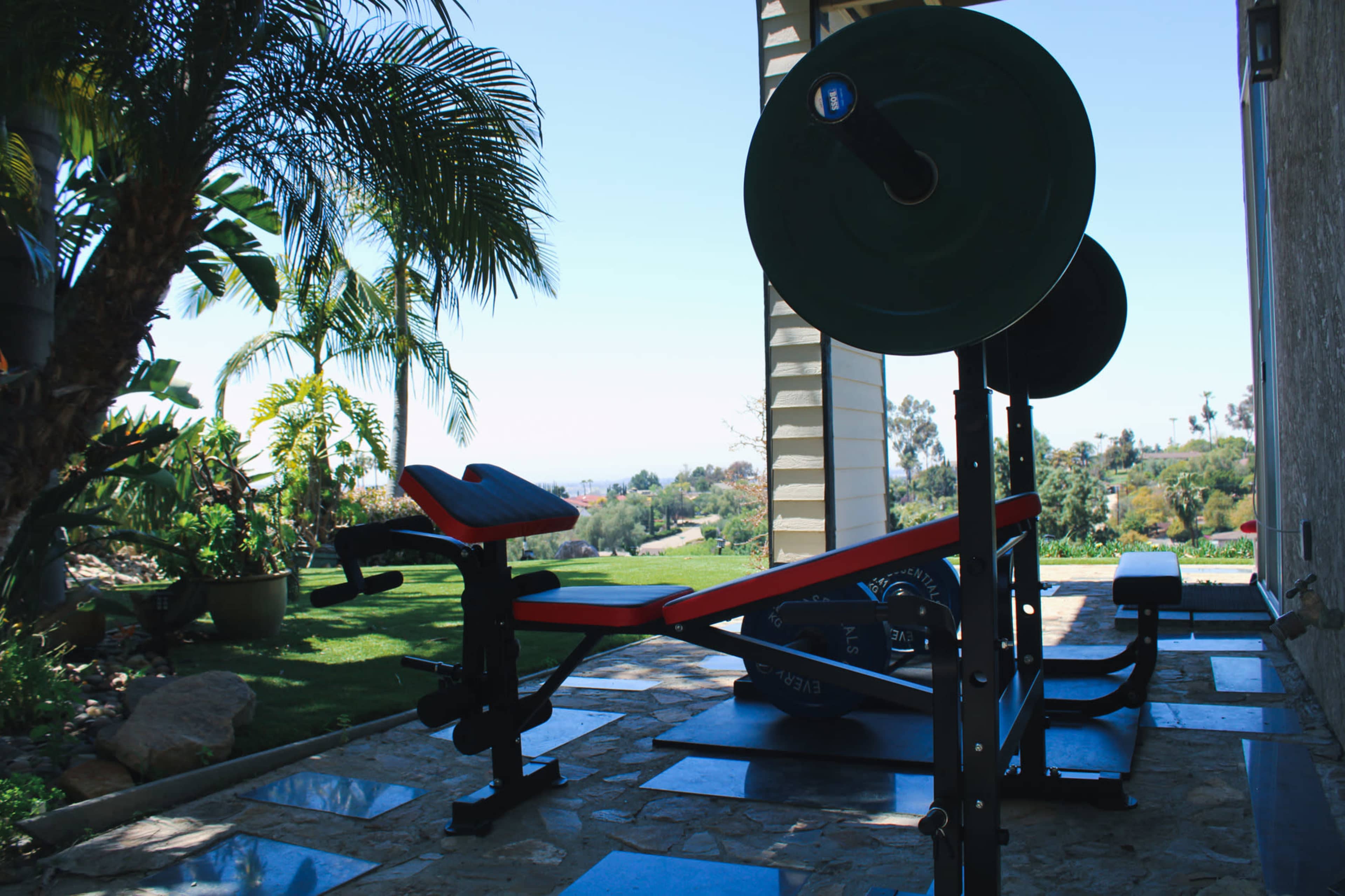 A weightlifting bench and barbell set positioned on a stone patio with a view of lush greenery in the background.