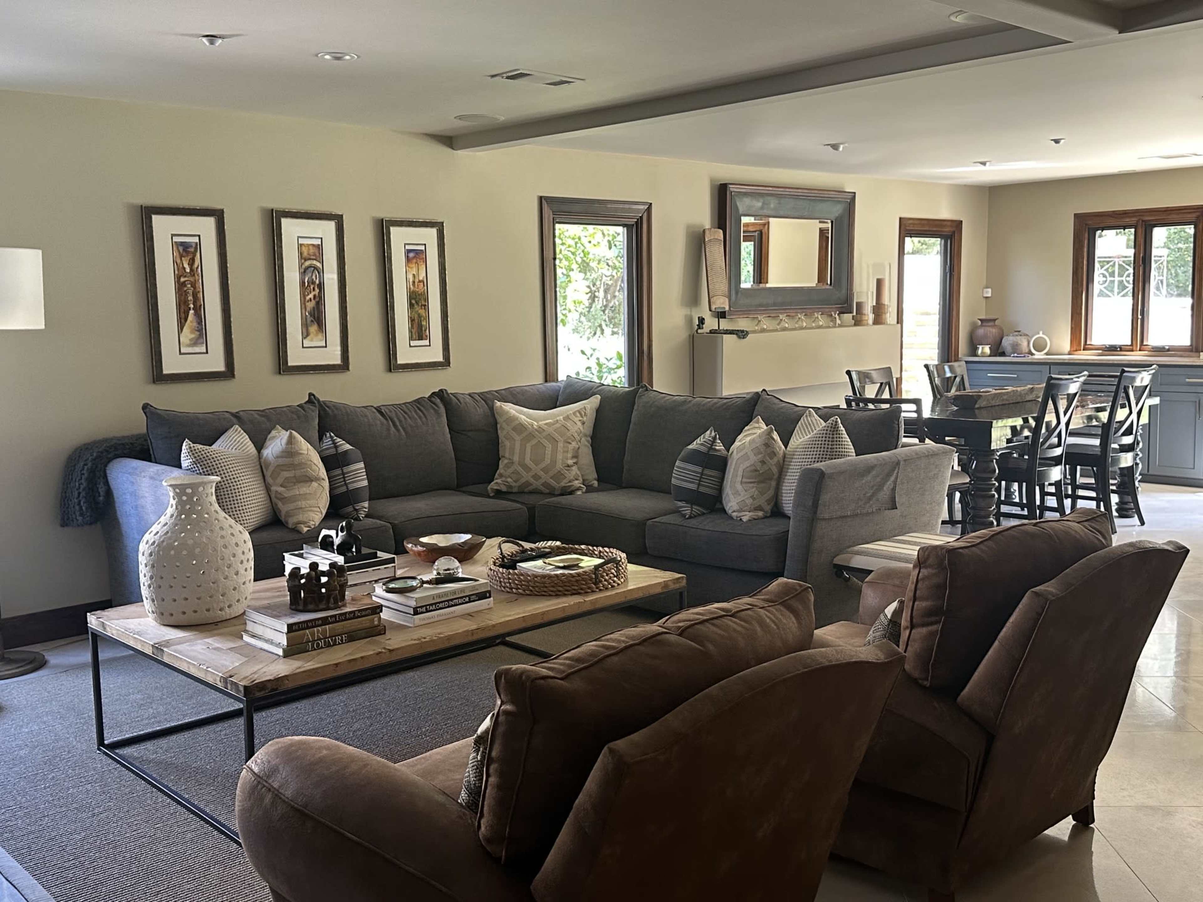 The image shows a modern living room with a large gray sectional sofa, a wooden coffee table, and decorative elements, including pillows and vases, set against a backdrop of windows and framed artwork.