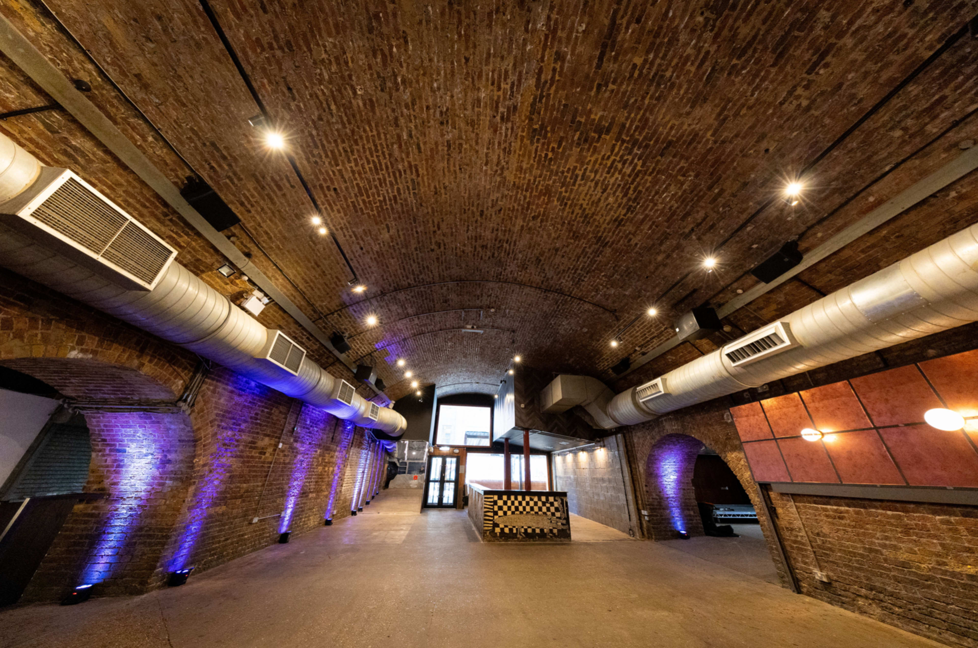 The image shows a spacious brick interior with an arched ceiling, featuring industrial-style lighting and visible air ducts along the walls.