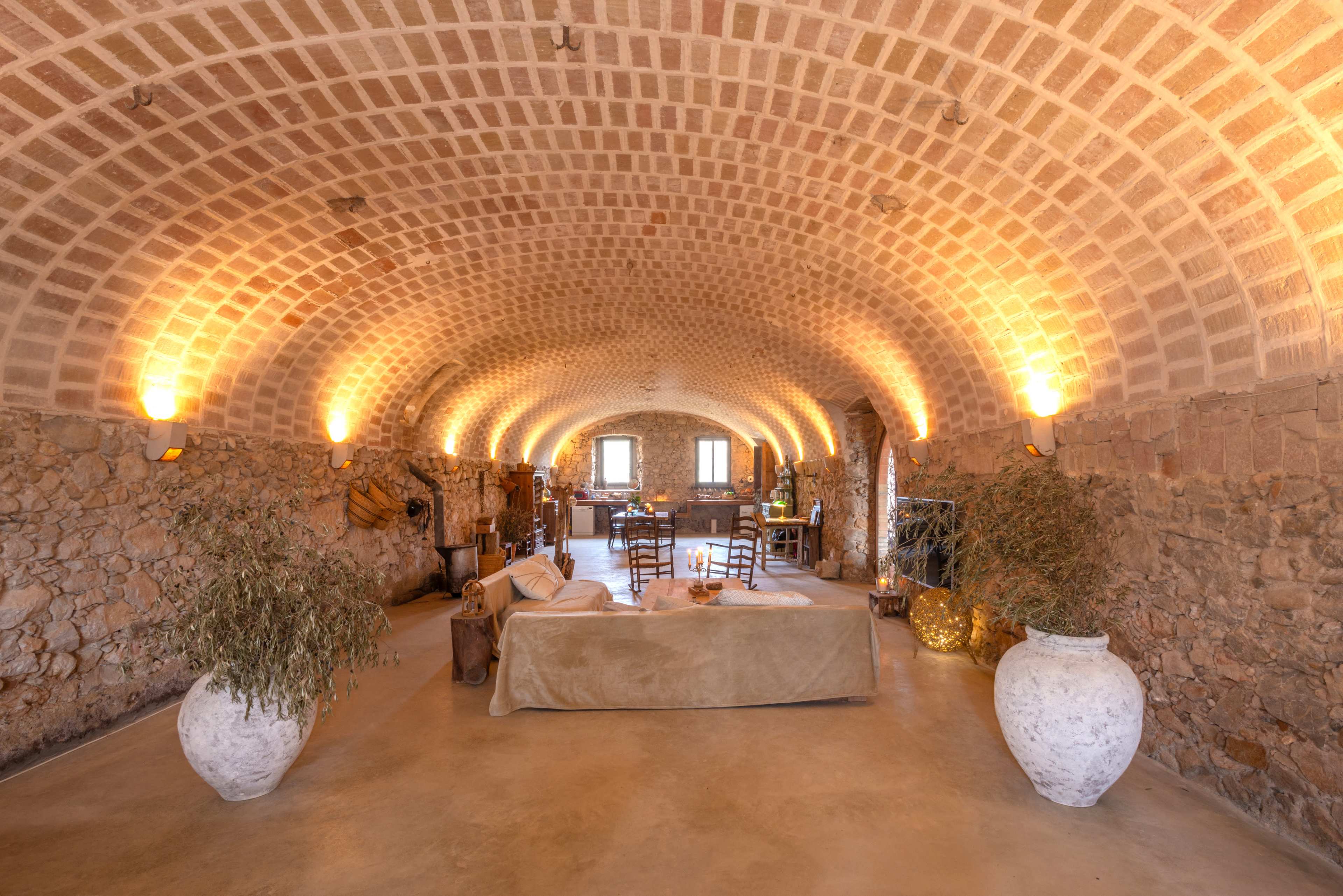 The image shows a spacious, semi-circular room with stone walls, arched ceilings, and warm lighting, featuring a sofa, decorative vases, and rustic furniture.