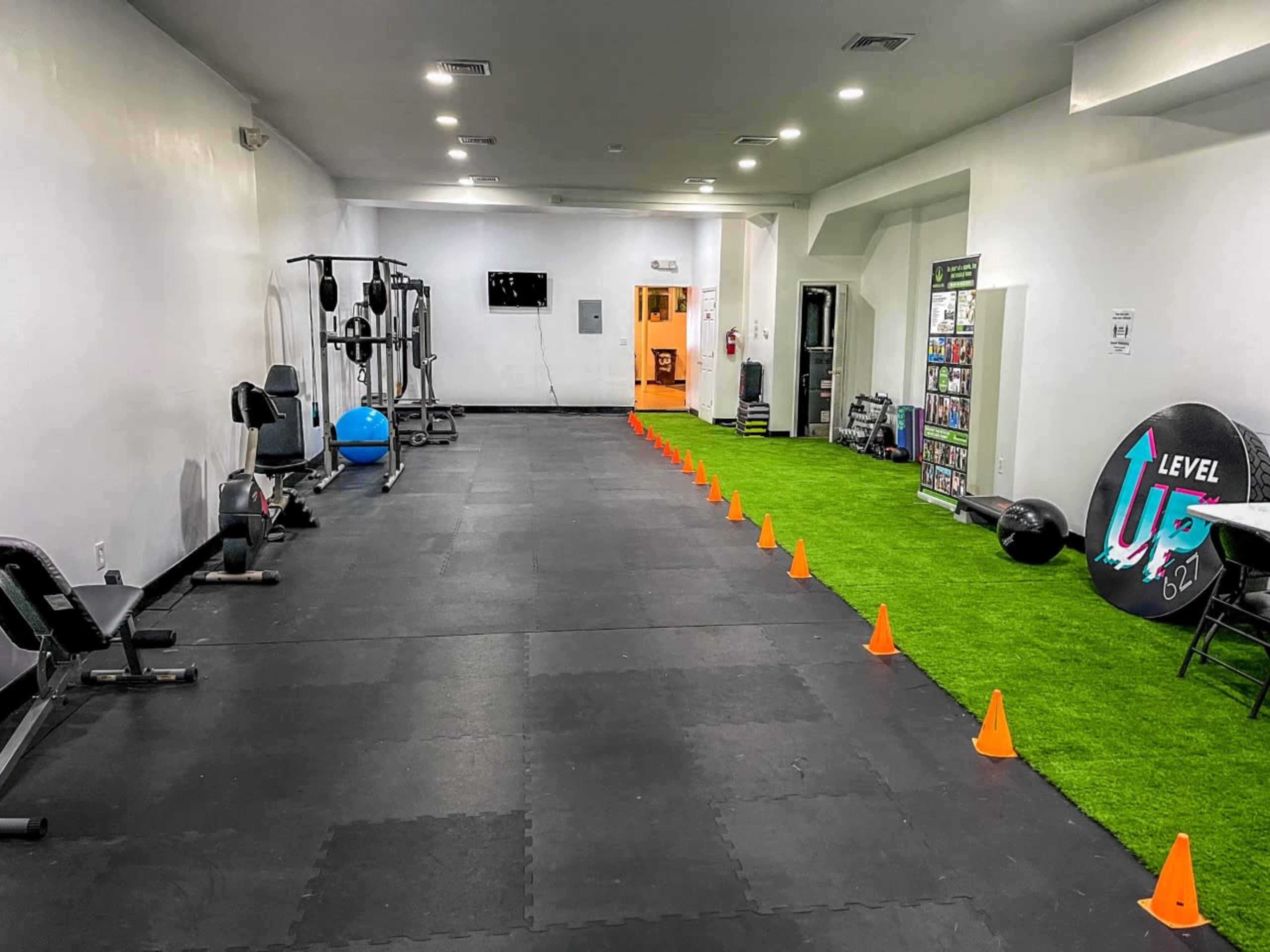 A gym interior with various exercise equipment on one side and a row of orange cones set on artificial grass along the floor.