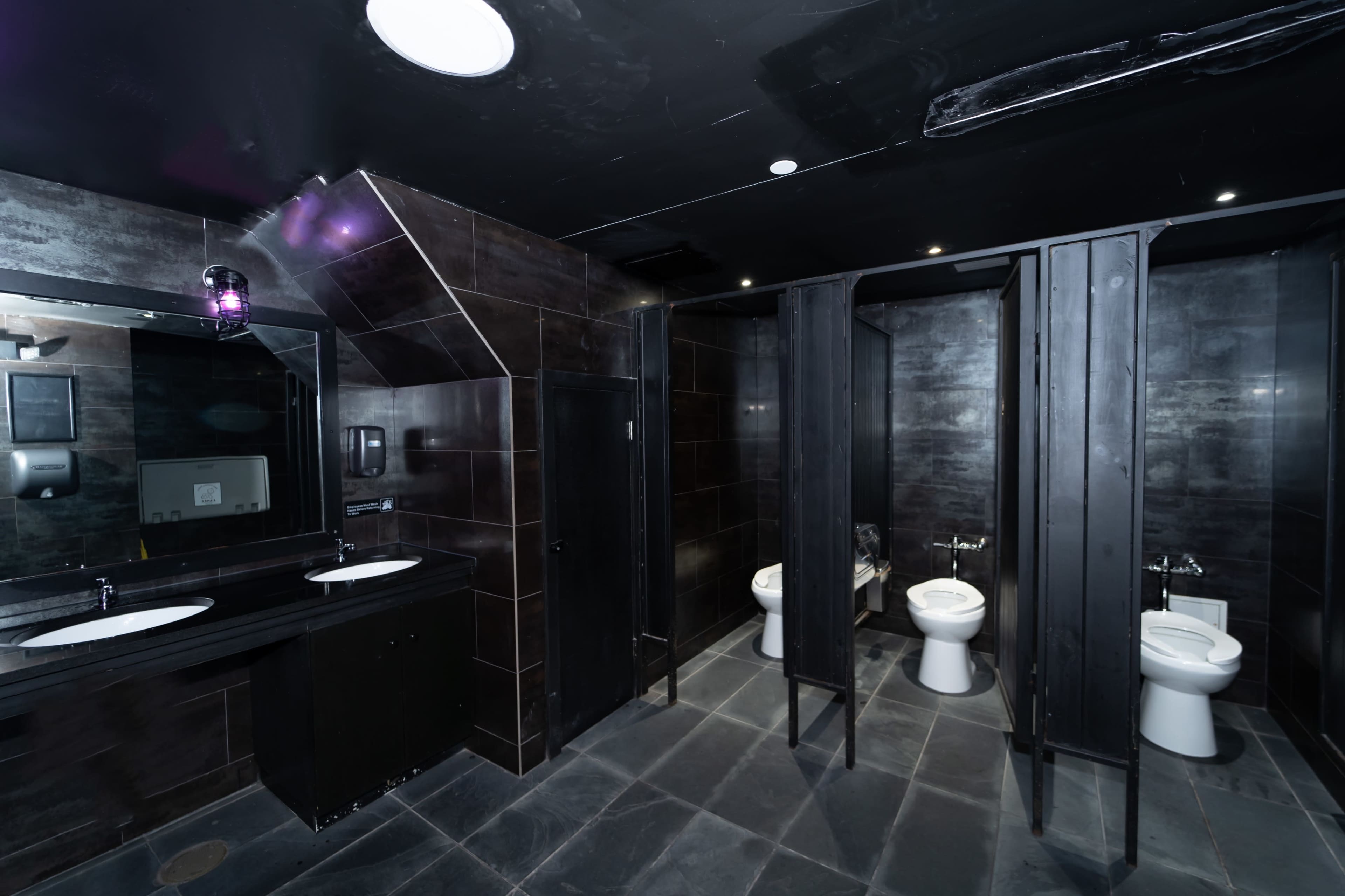 A modern restroom with two stalls, a row of sinks, and dark tiled walls and floors.
