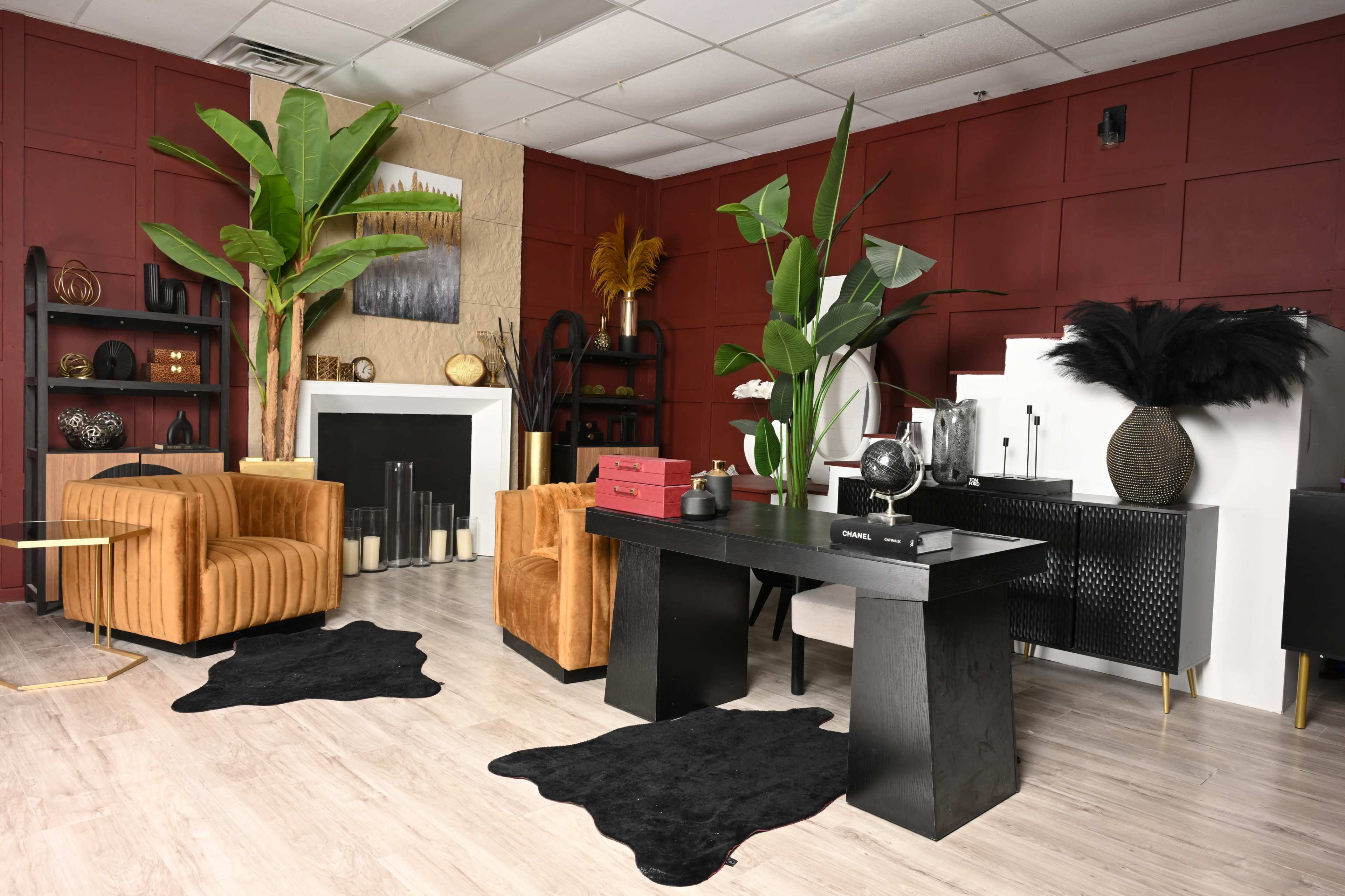 The room features a modern office setup with two orange velvet chairs, a black desk, and decorative plants, accented by a red and beige color scheme.