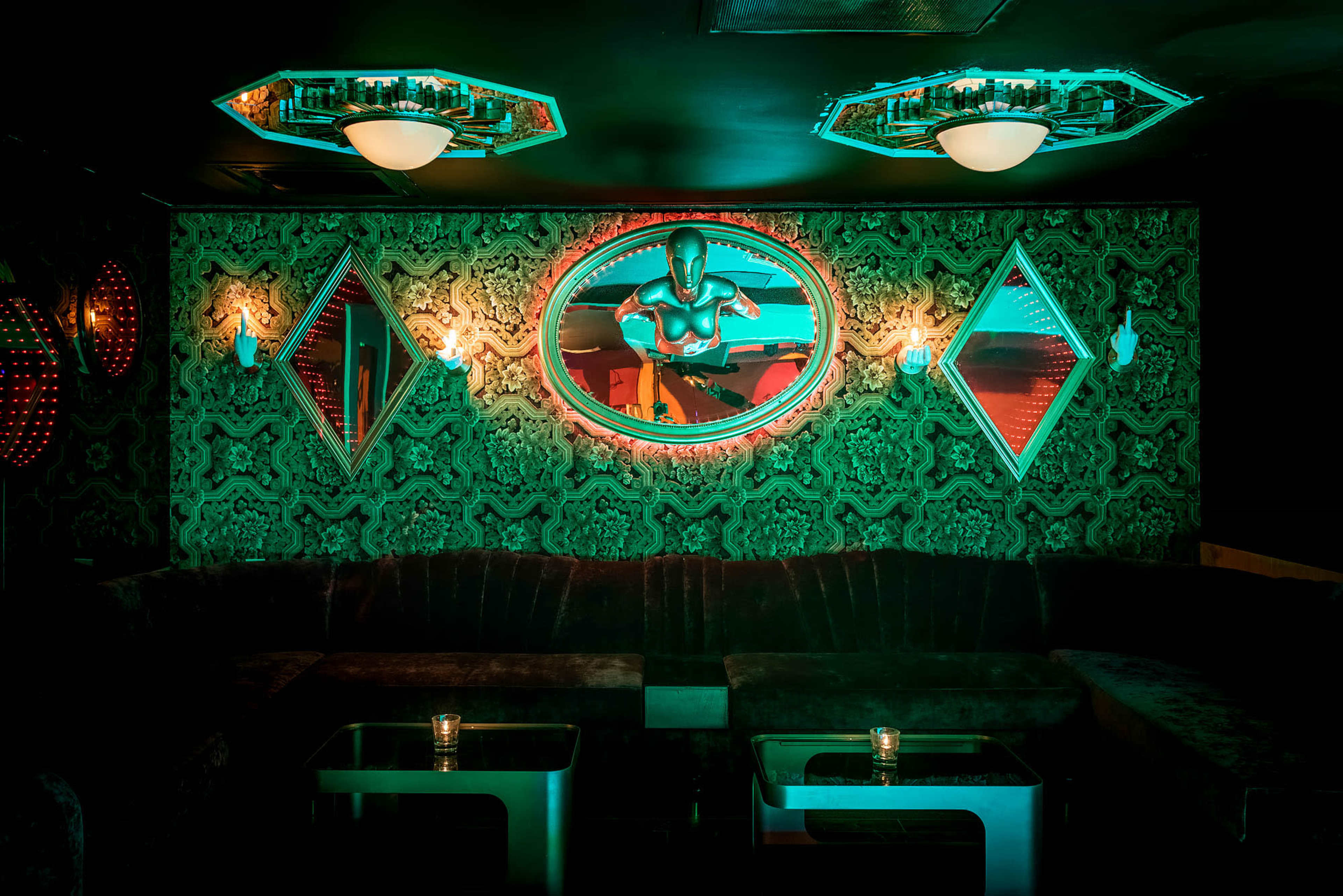 A dimly lit lounge features a plush velvet seating area, decorative mirrors, and green-tinted lighting highlighting the walls.