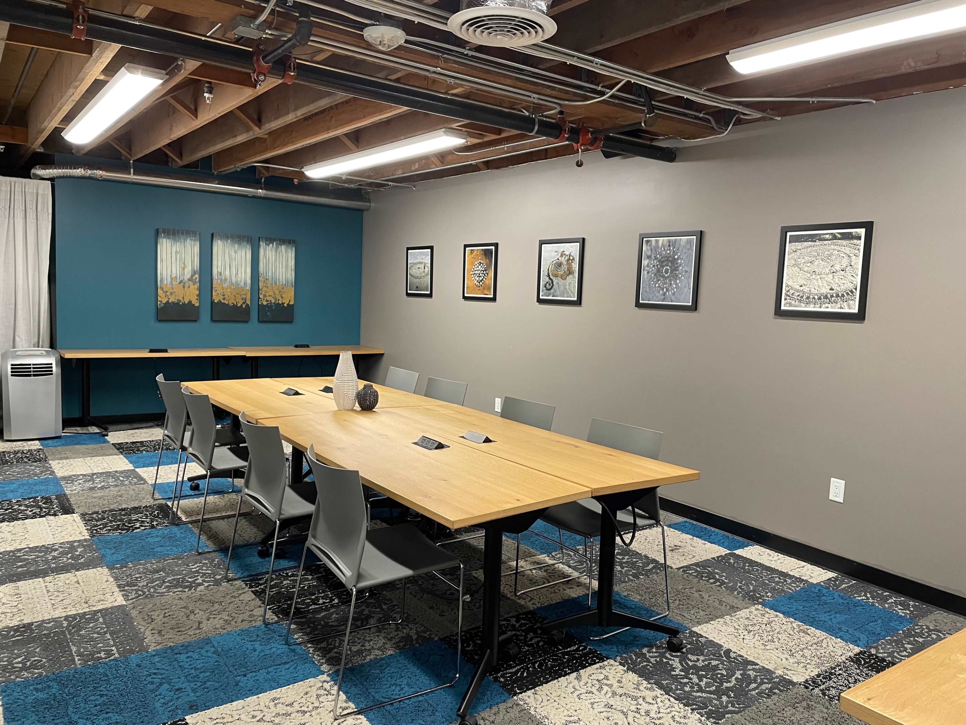 A modern conference room with a long wooden table surrounded by gray chairs, decorated with framed artwork on the walls and featuring a large air conditioning unit in the corner.