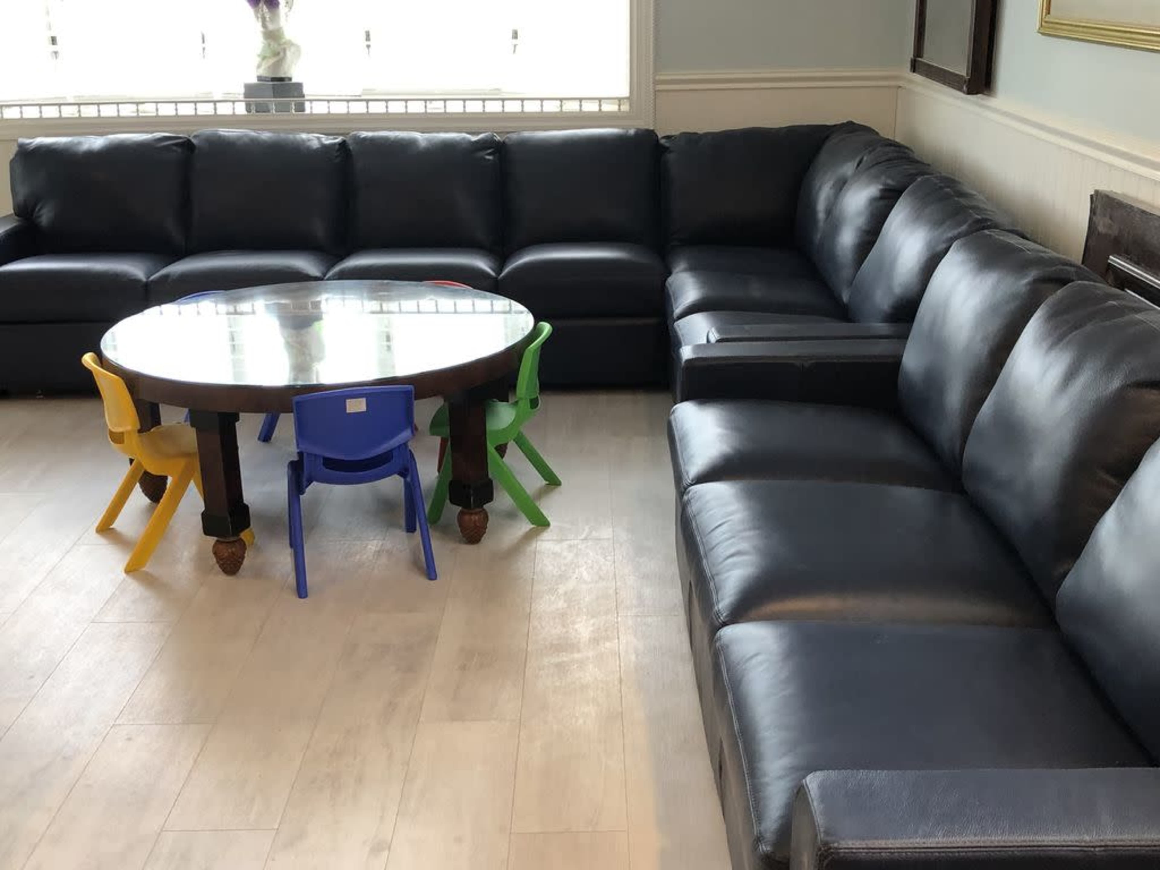 A large black leather sectional sofa surrounding a round wooden table with four colorful children's chairs.