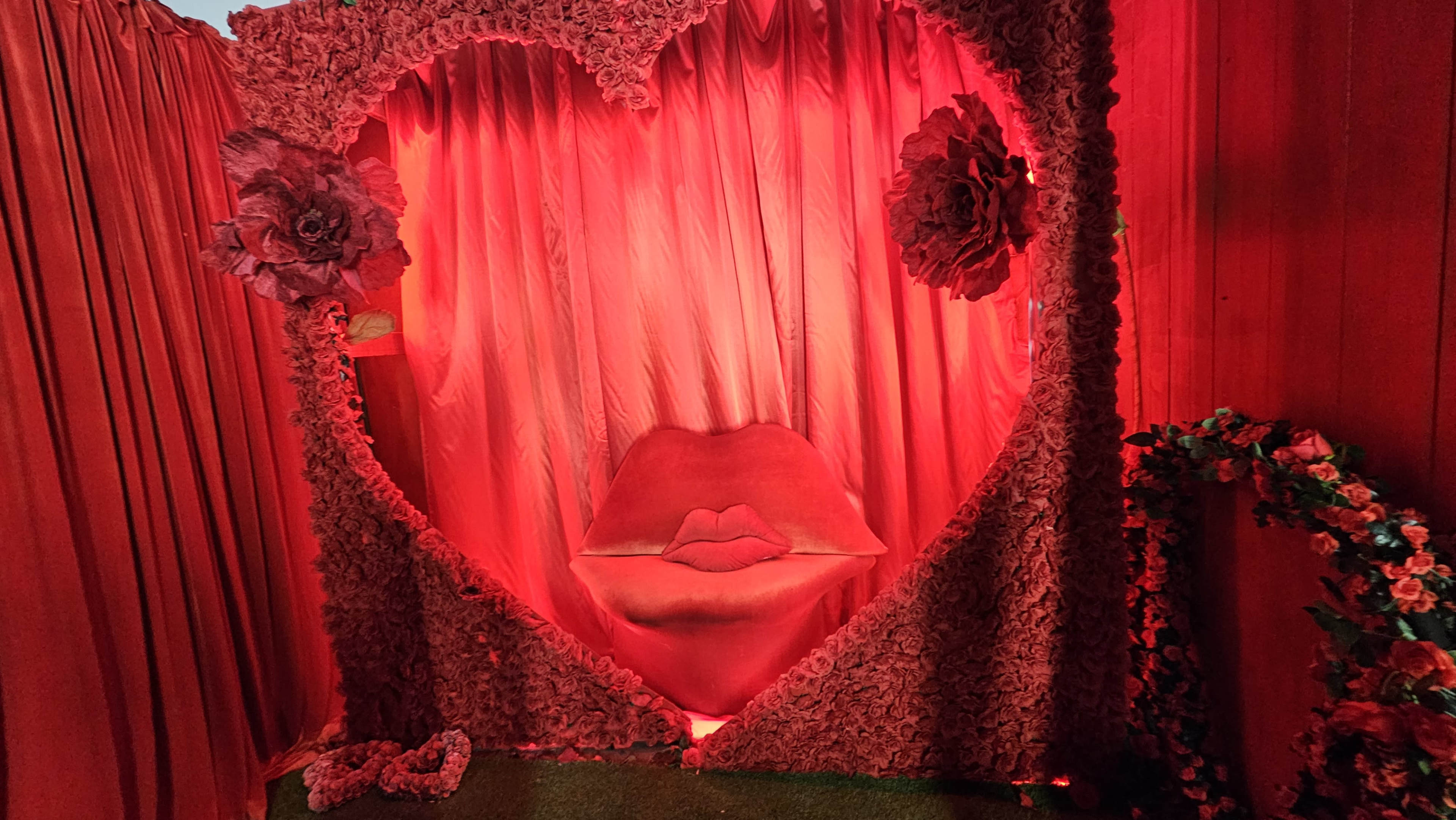 A heart-shaped backdrop adorned with red flowers and a sofa designed to resemble lips, illuminated by red lighting.