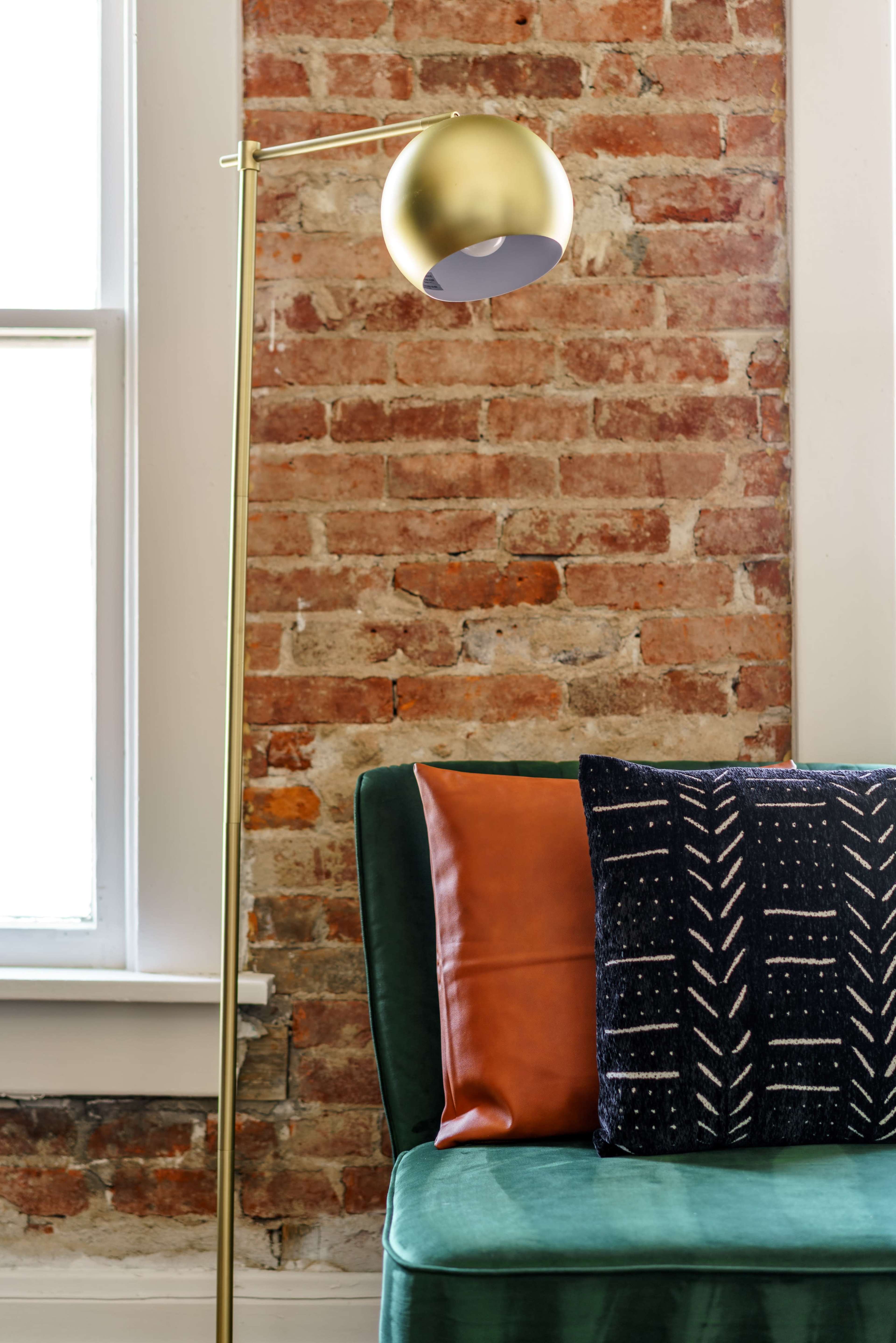 A gold floor lamp stands beside a green couch adorned with a brown leather pillow and a patterned black pillow, all set against a brick wall.