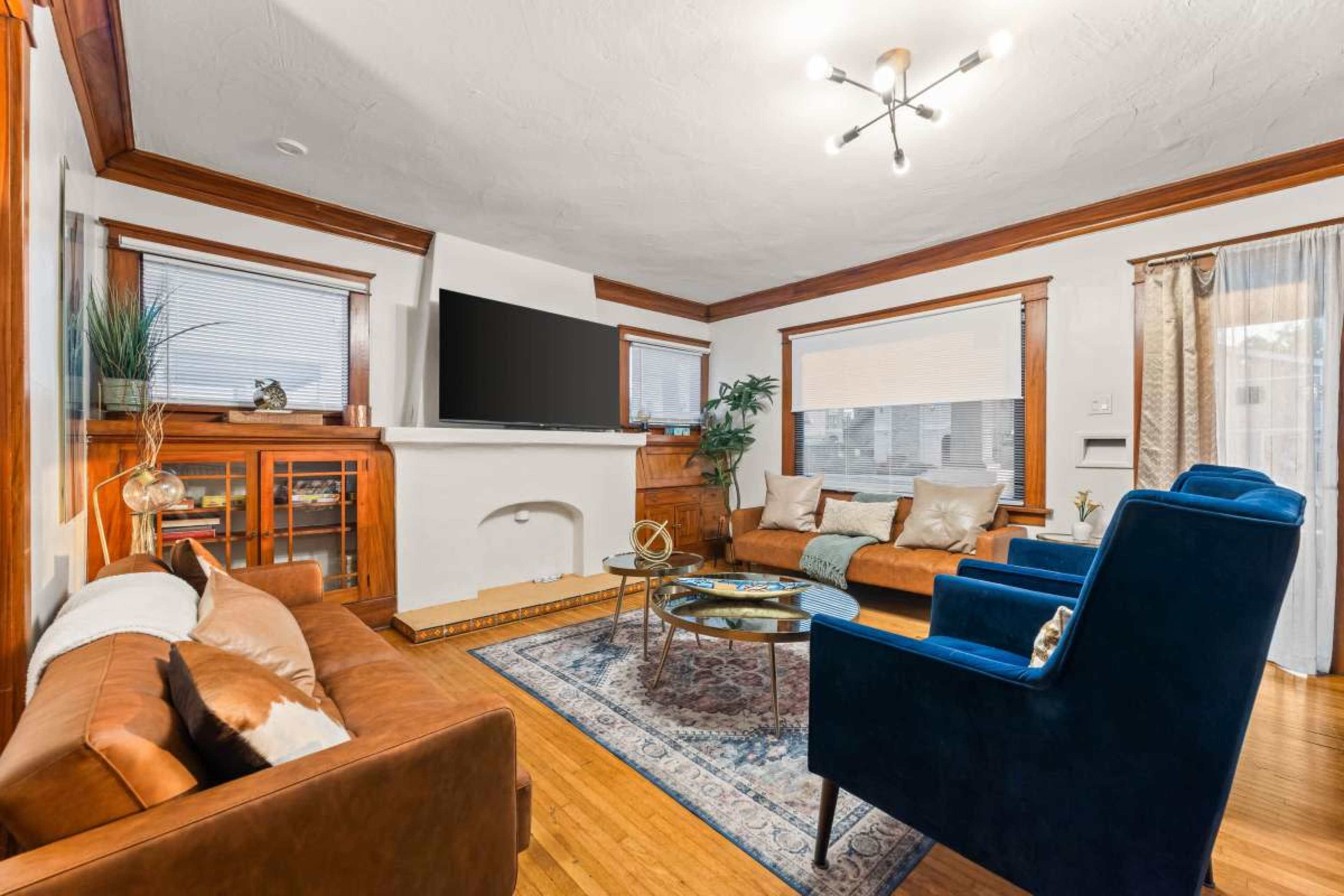 The image shows a cozy living room with two brown leather sofas, a blue upholstered armchair, a coffee table, and a television mounted above a white fireplace.
