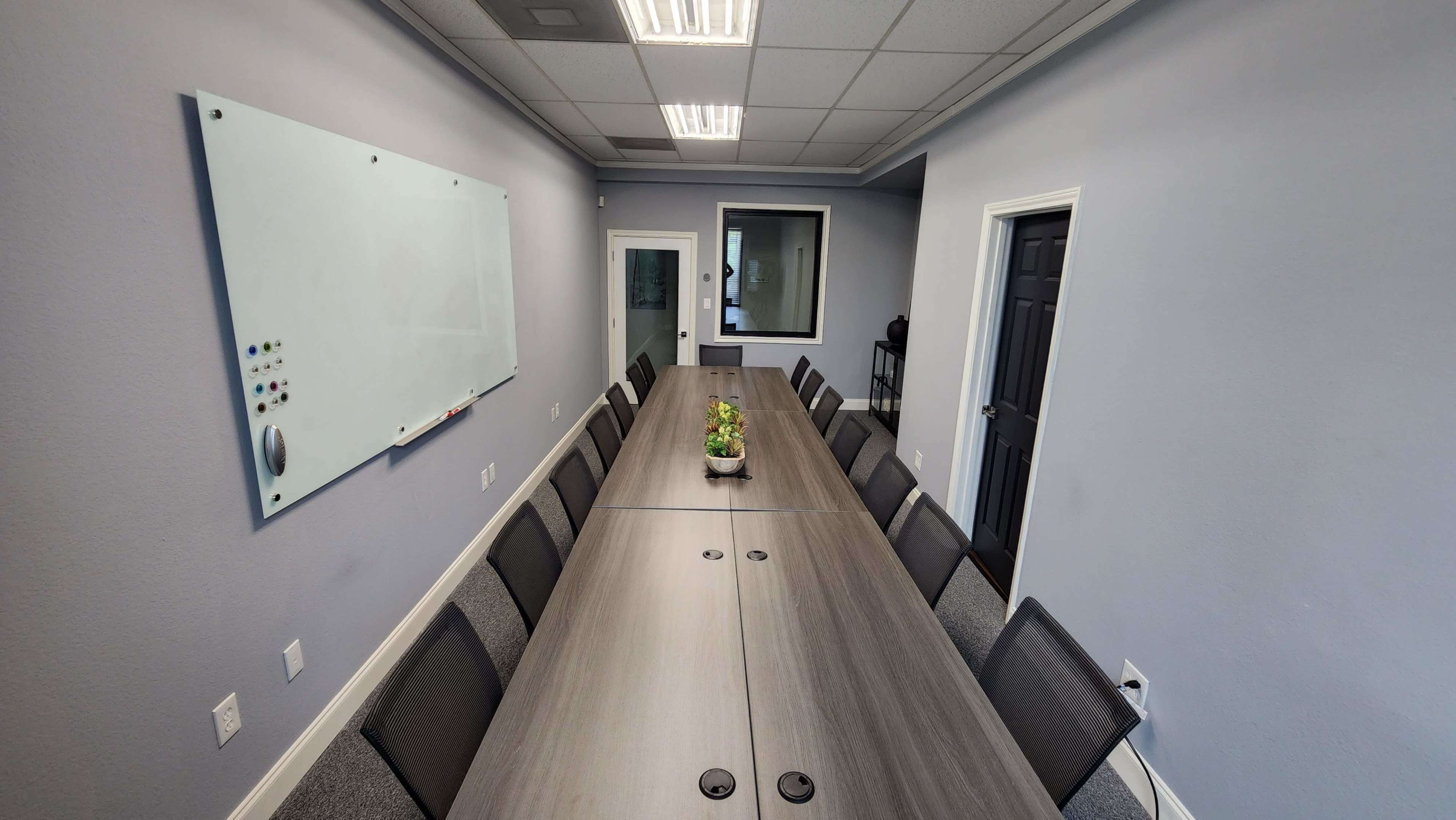 A long conference table is set up in a meeting room, flanked by several chairs, with a plant centerpiece and a whiteboard on the wall.
