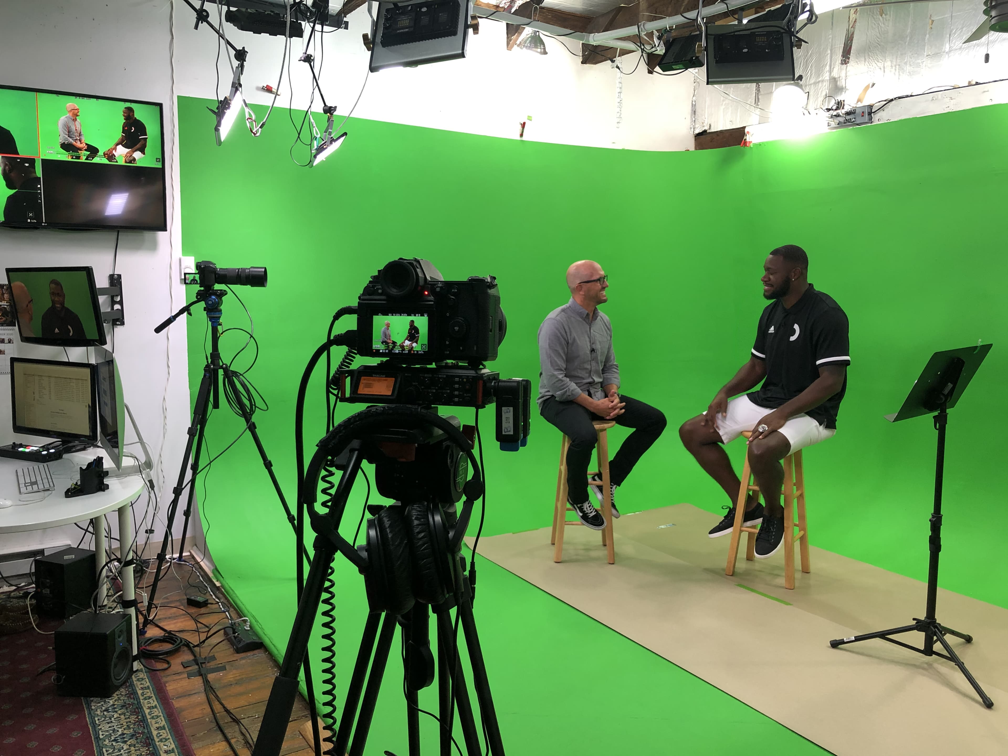 Two men sitting on stools in front of a green screen, with cameras and a monitor set up for filming in a studio.