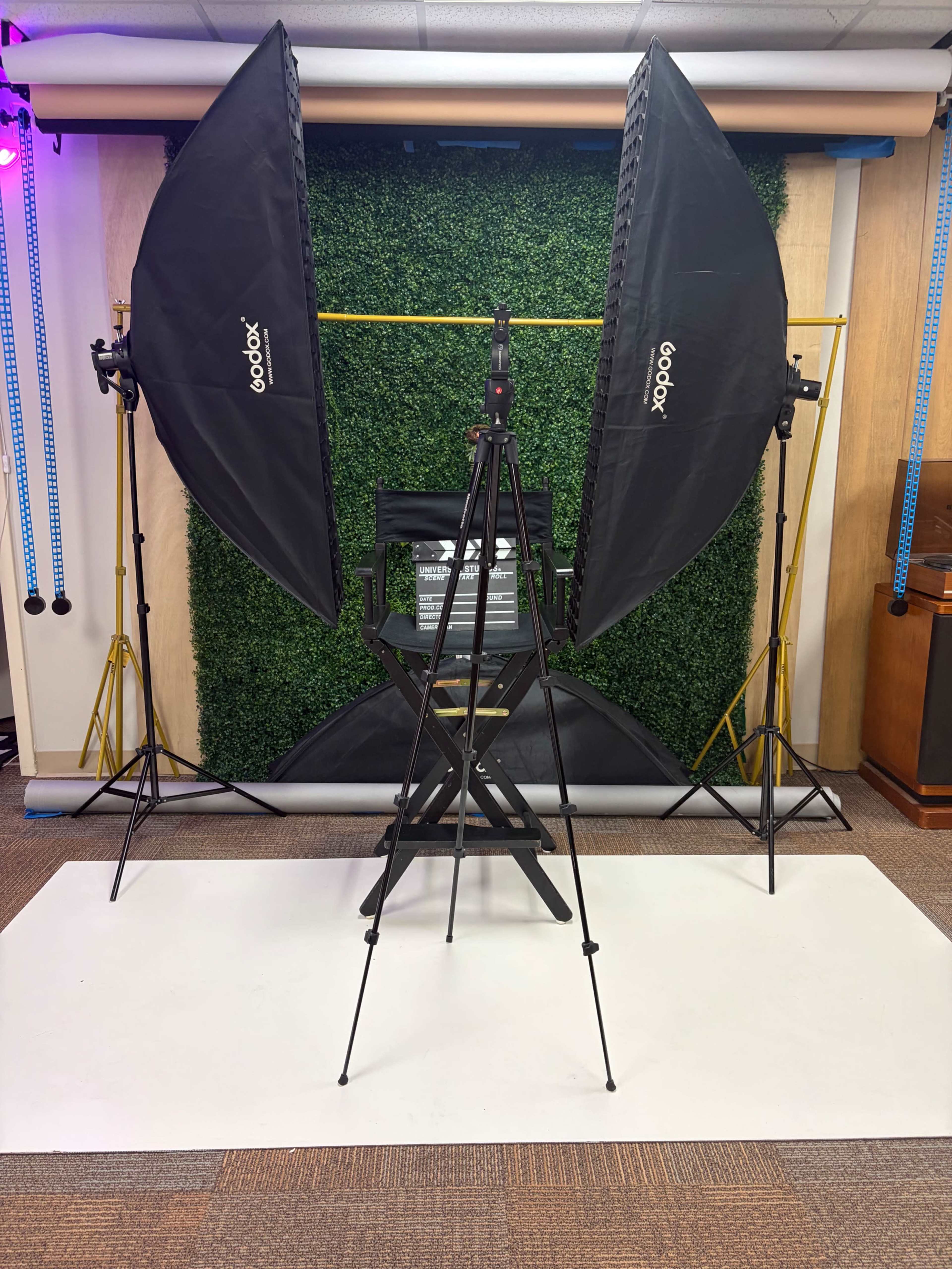The image shows a photo studio setup featuring two large softboxes on tripods, a director's chair in front of a green backdrop, and a white floor covering.