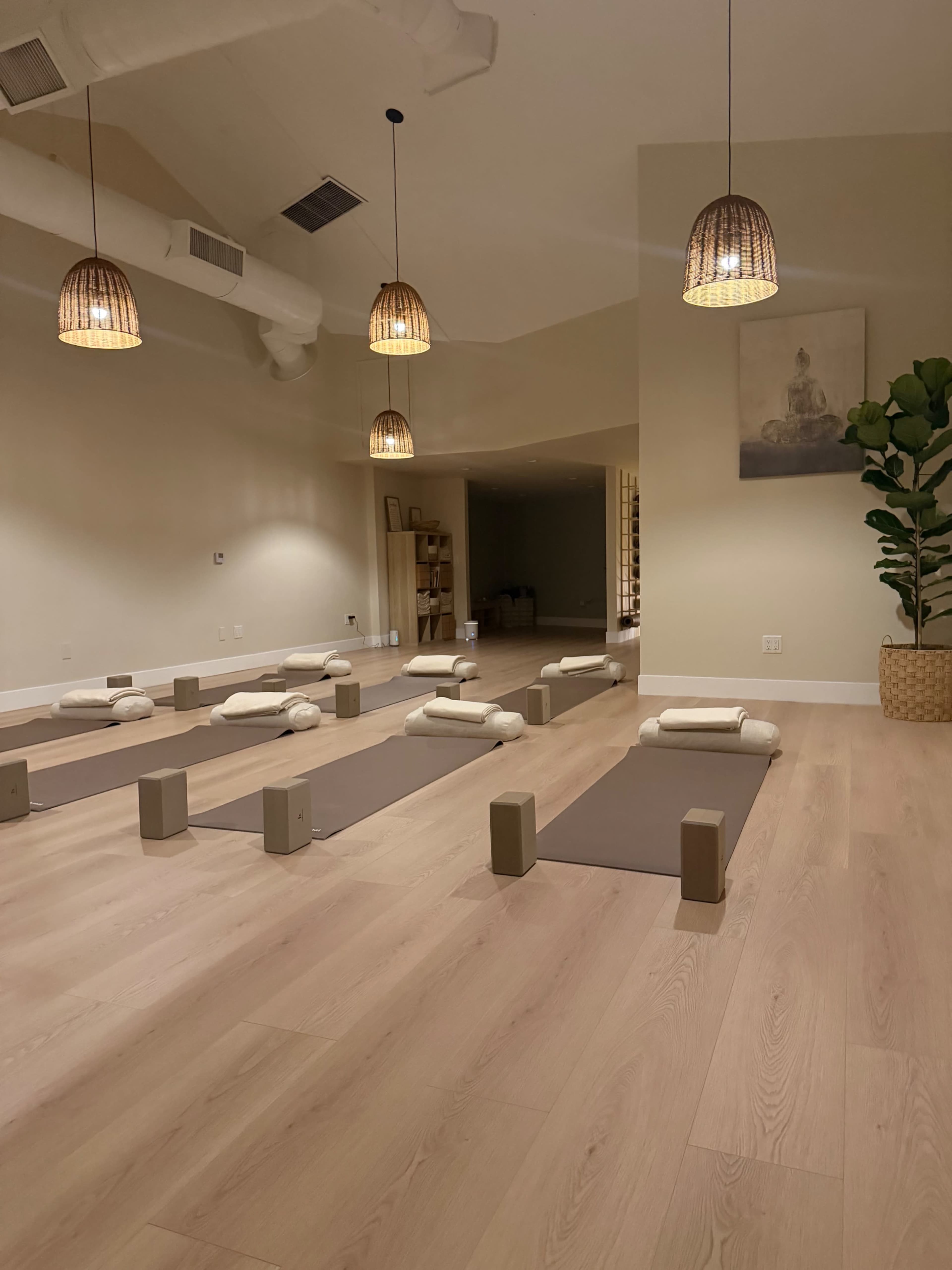 The image shows a serene yoga studio with rolled mats and props arranged neatly on a wooden floor, illuminated by pendant lights.