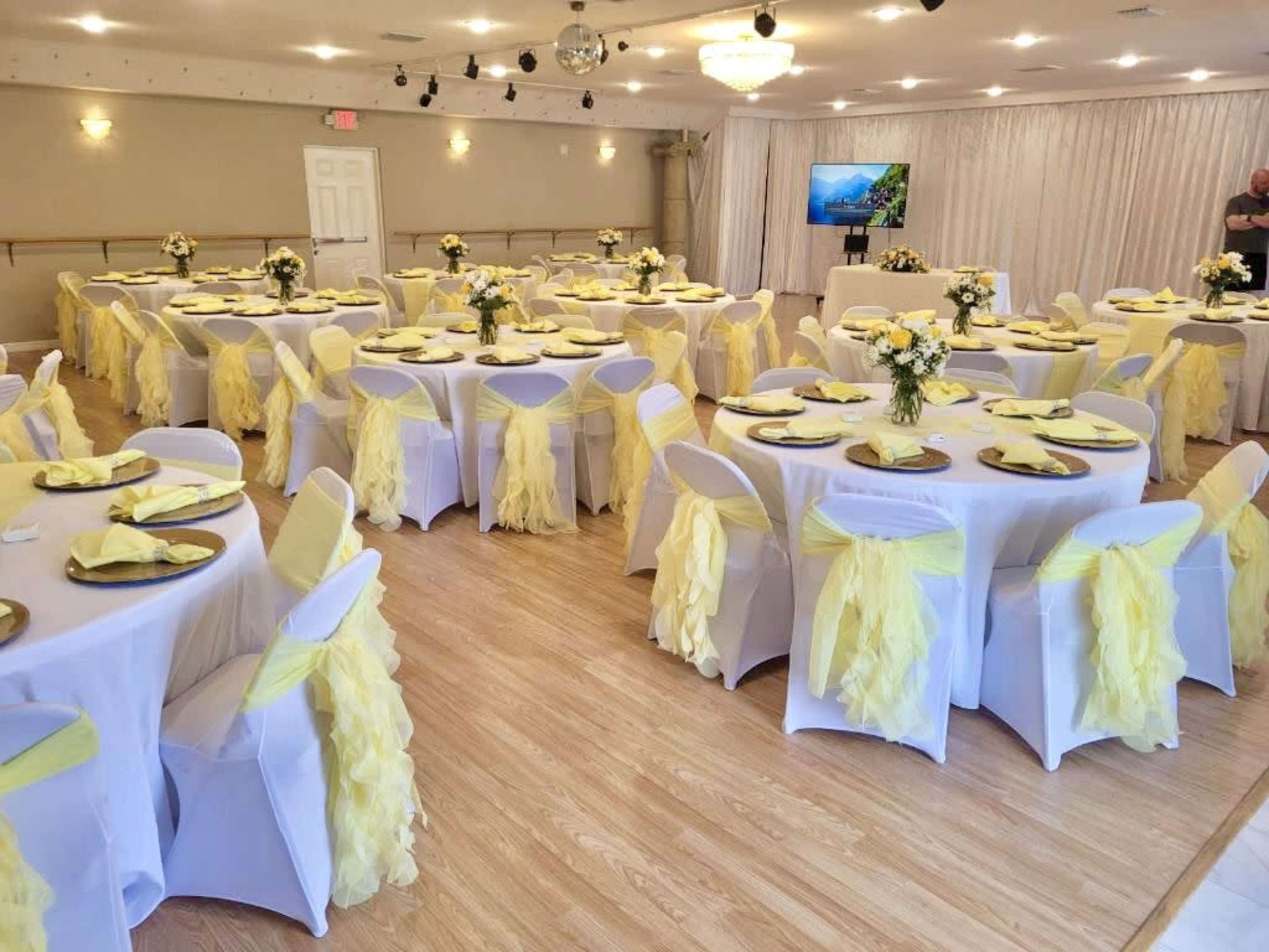 A banquet hall set up for an event with round tables covered in white tablecloths and decorated with yellow-tinted centerpieces, surrounded by chairs draped in yellow fabric.