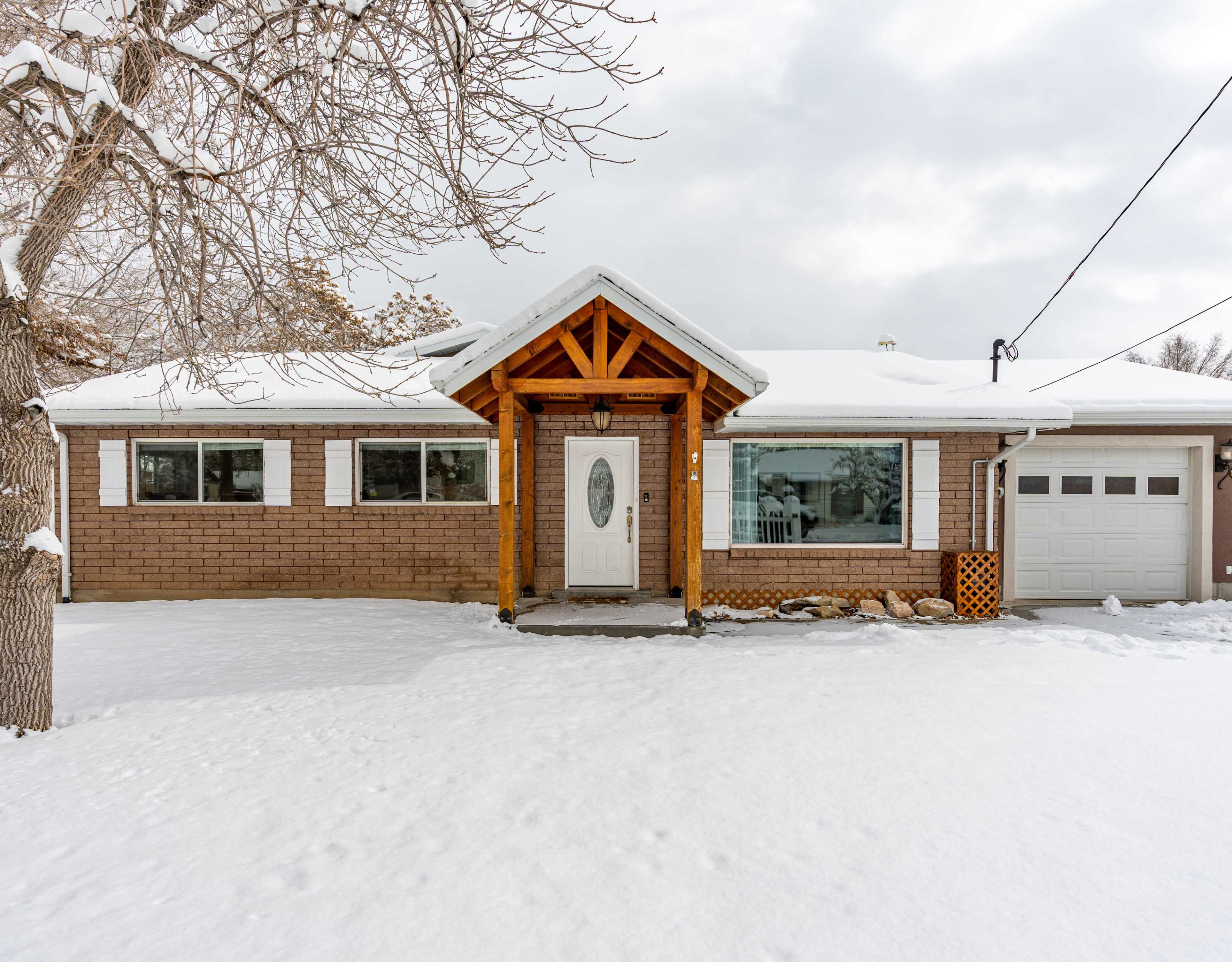 A single-story brick house with a wooden porch structure is surrounded by snow, featuring large front windows and an attached garage.