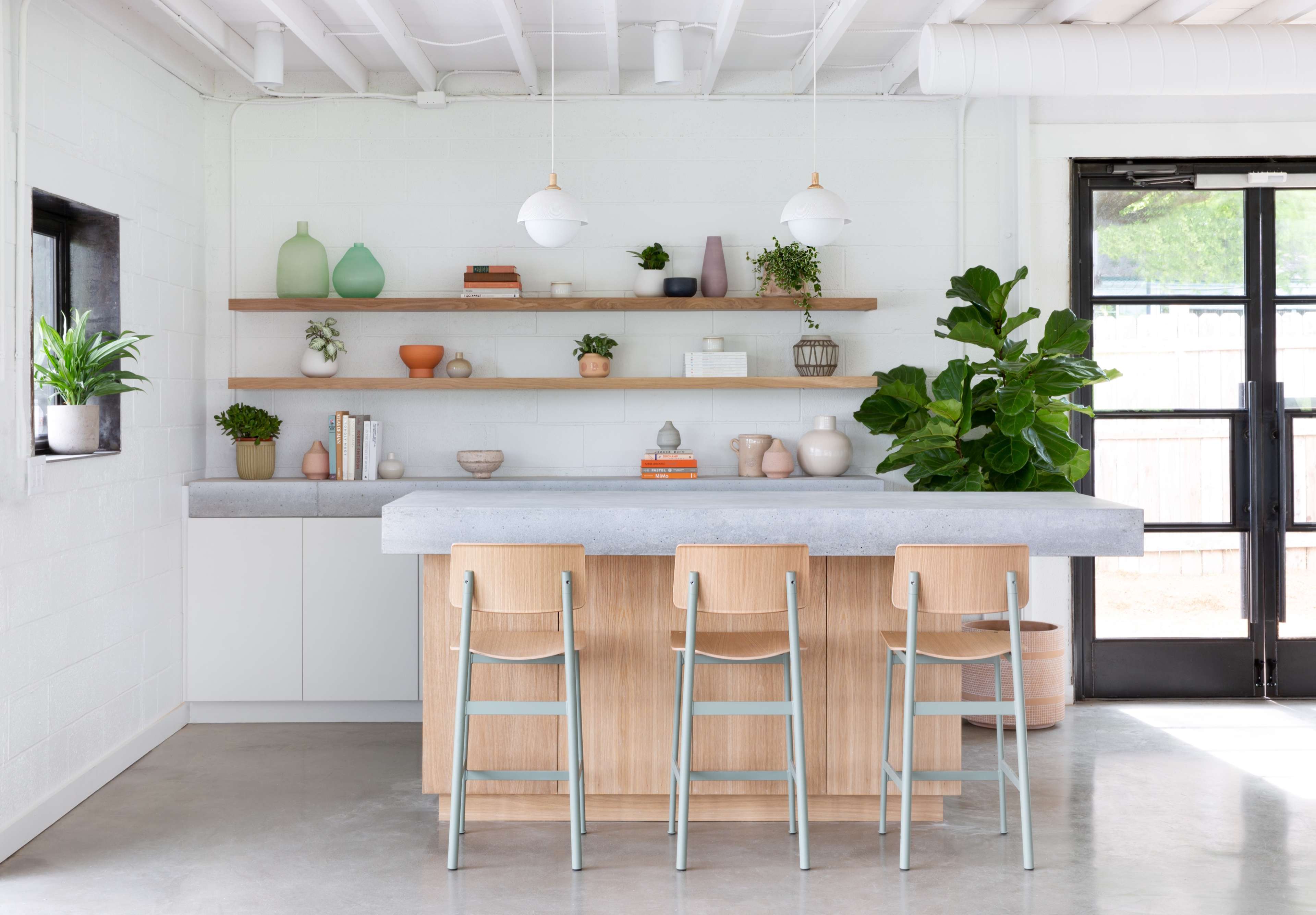 A modern kitchen features a concrete island with wooden stools, wall shelves displaying various decorative items, and plants in pots.