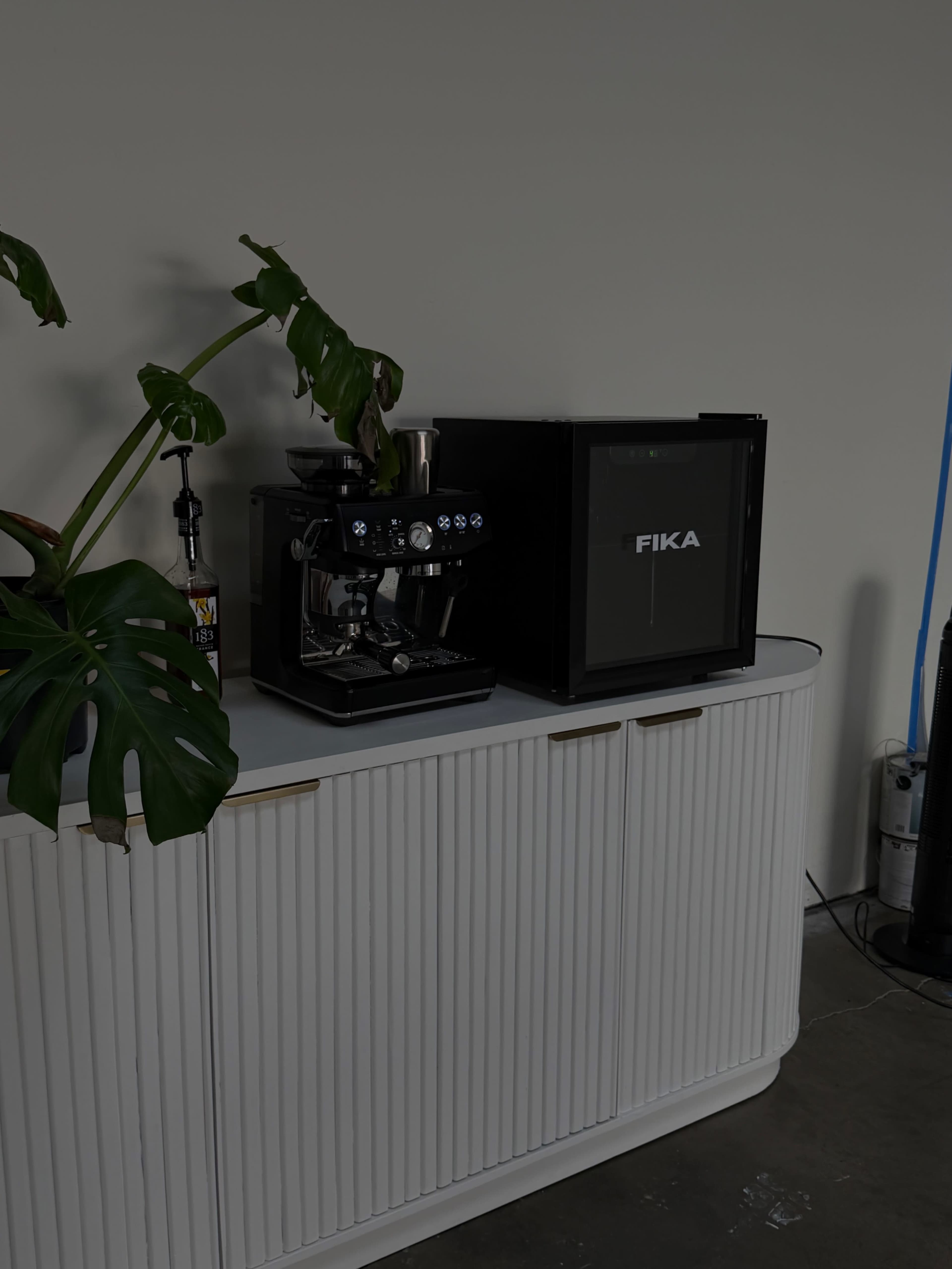 A coffee machine and a FIKA mini fridge are placed on top of a white cabinet with ribbed detailing, accompanied by a large green plant.