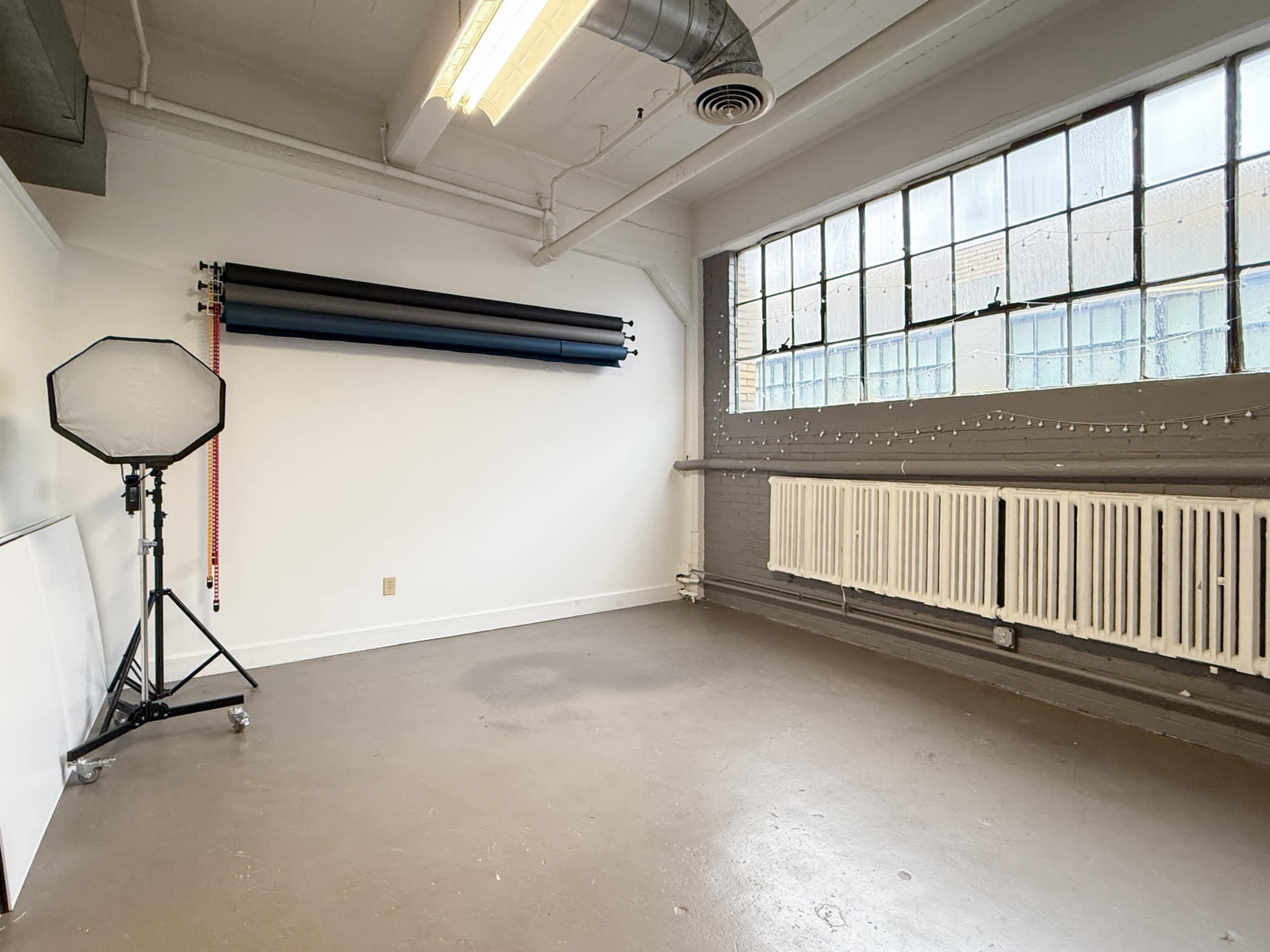 The image shows a spacious studio with a large window, a backdrop support system, and a radiator along the wall.