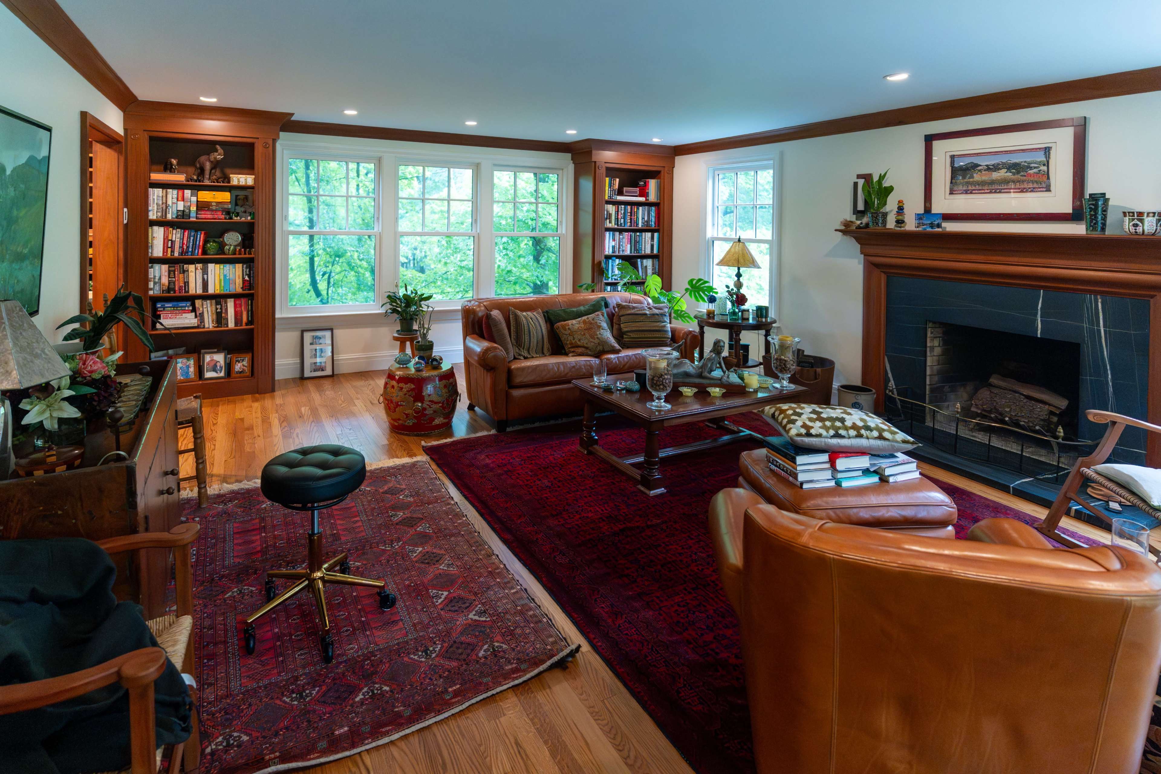 The image depicts a cozy living room with a fireplace, wooden bookshelves, and large windows overlooking greenery.