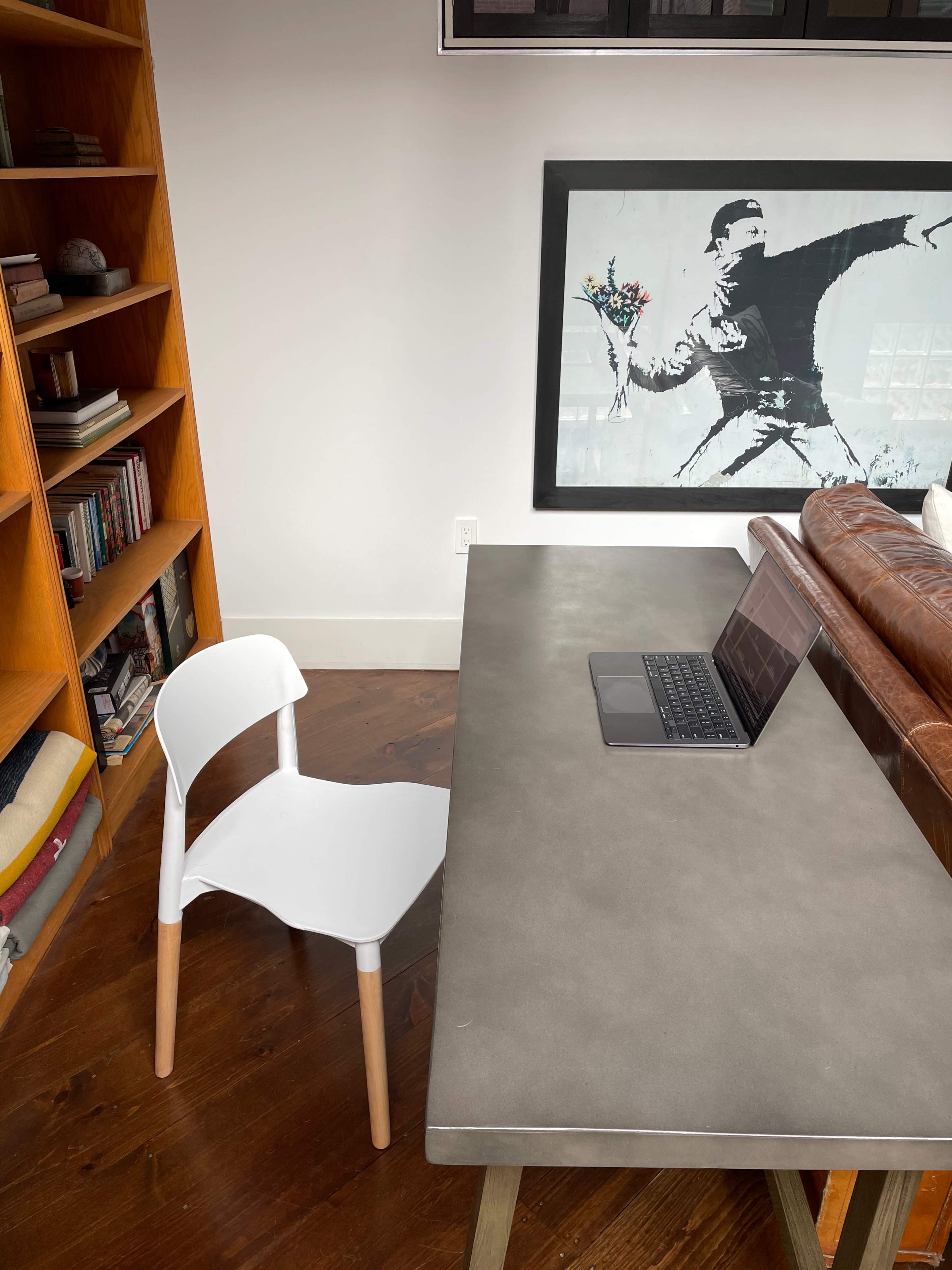 A minimalist desk with a laptop and an empty white chair is positioned near a bookshelf and a framed artwork on the wall.