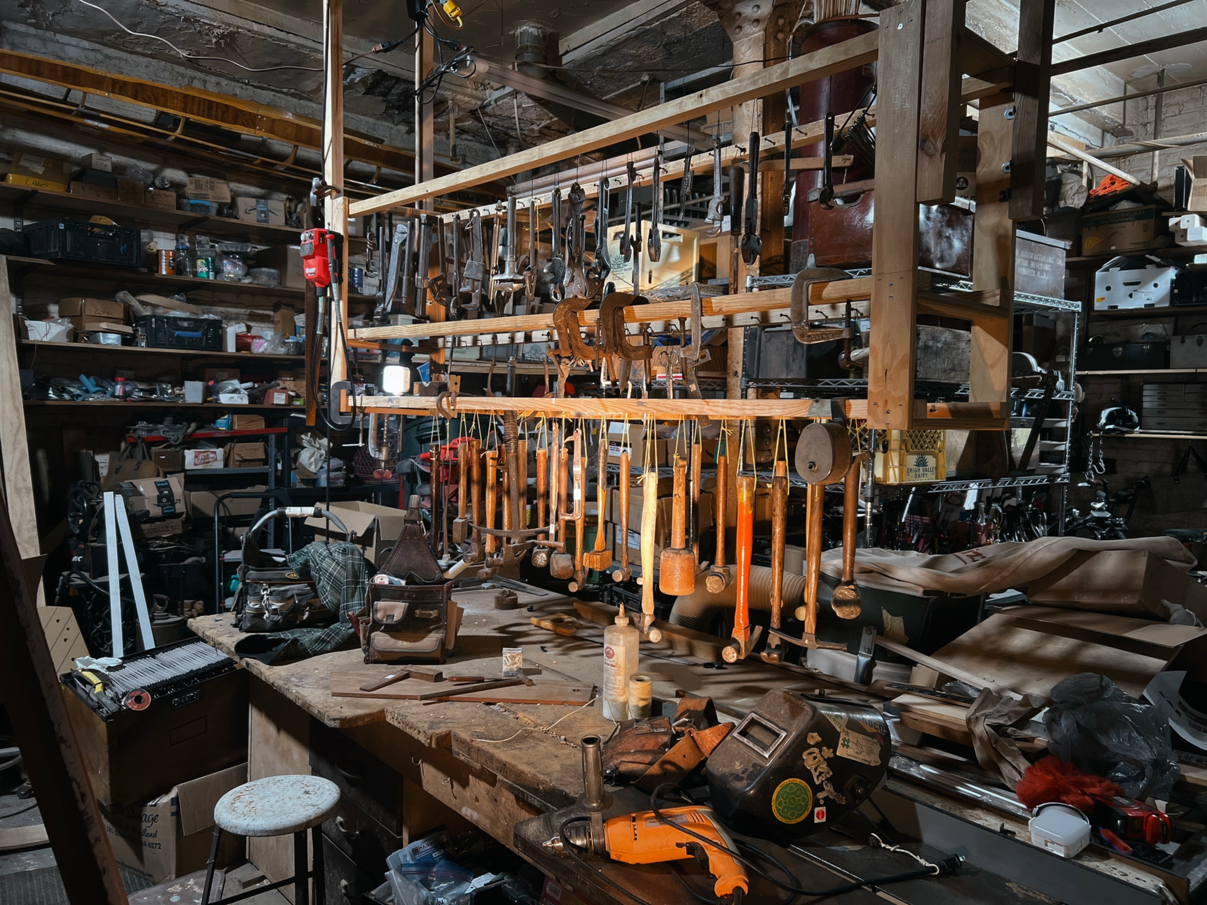 A cluttered workshop is filled with various tools hanging from a wooden rack and scattered across a workbench.