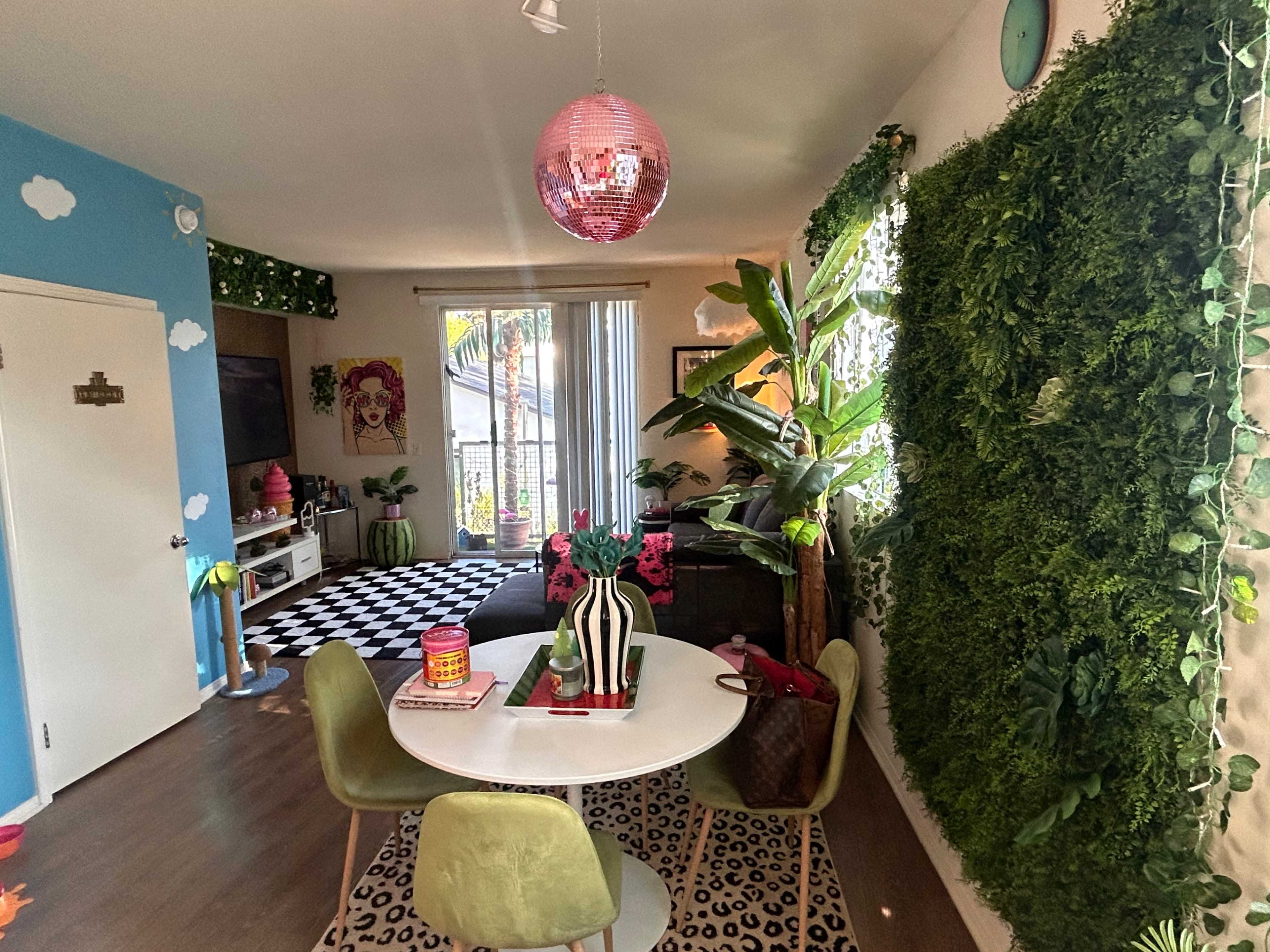 A modern living space features a round dining table with green chairs, a pink disco ball overhead, and lush greenery on the walls.