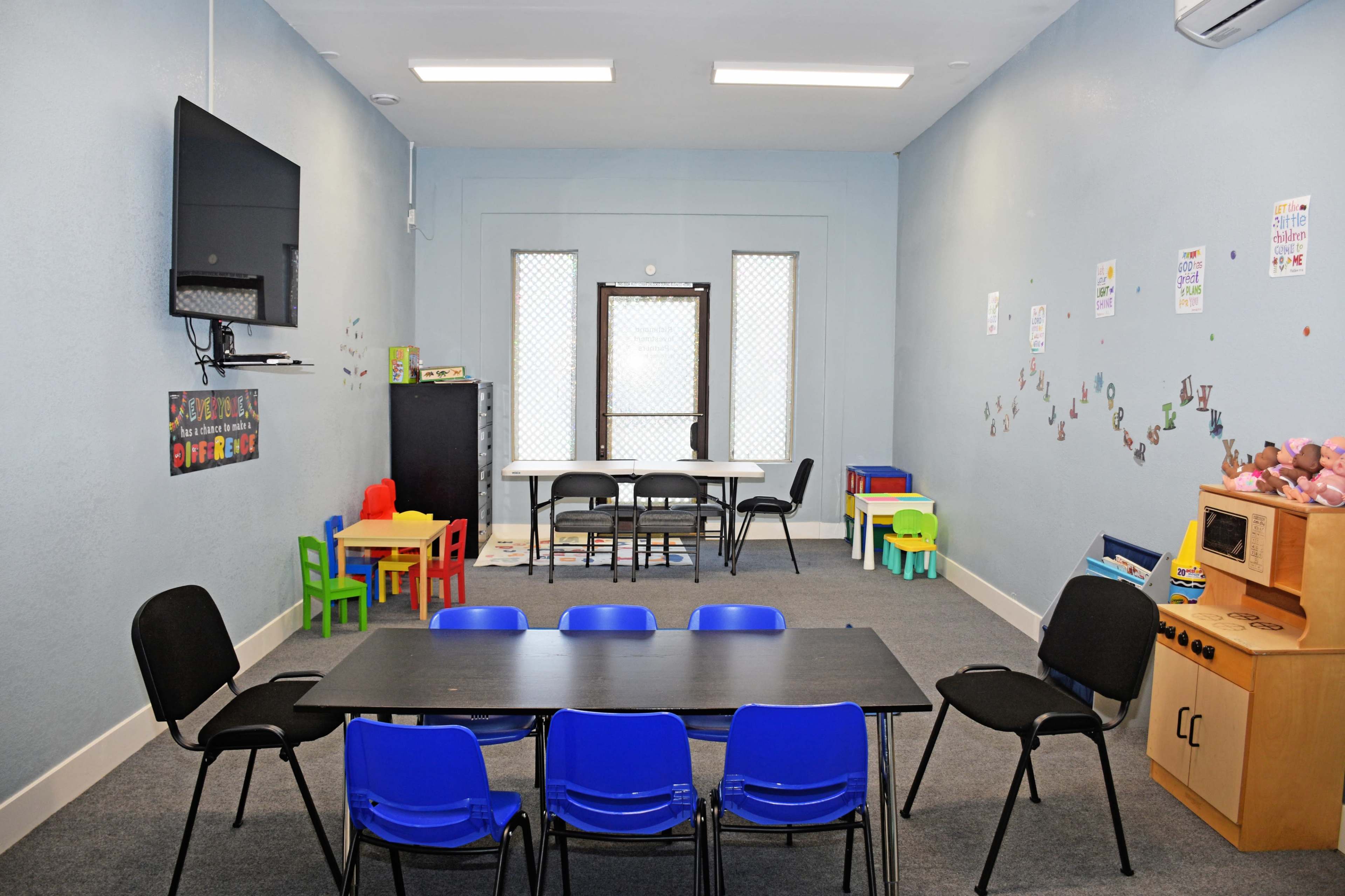 A brightly colored classroom features multiple tables and chairs, a TV on the wall, and educational decorations.