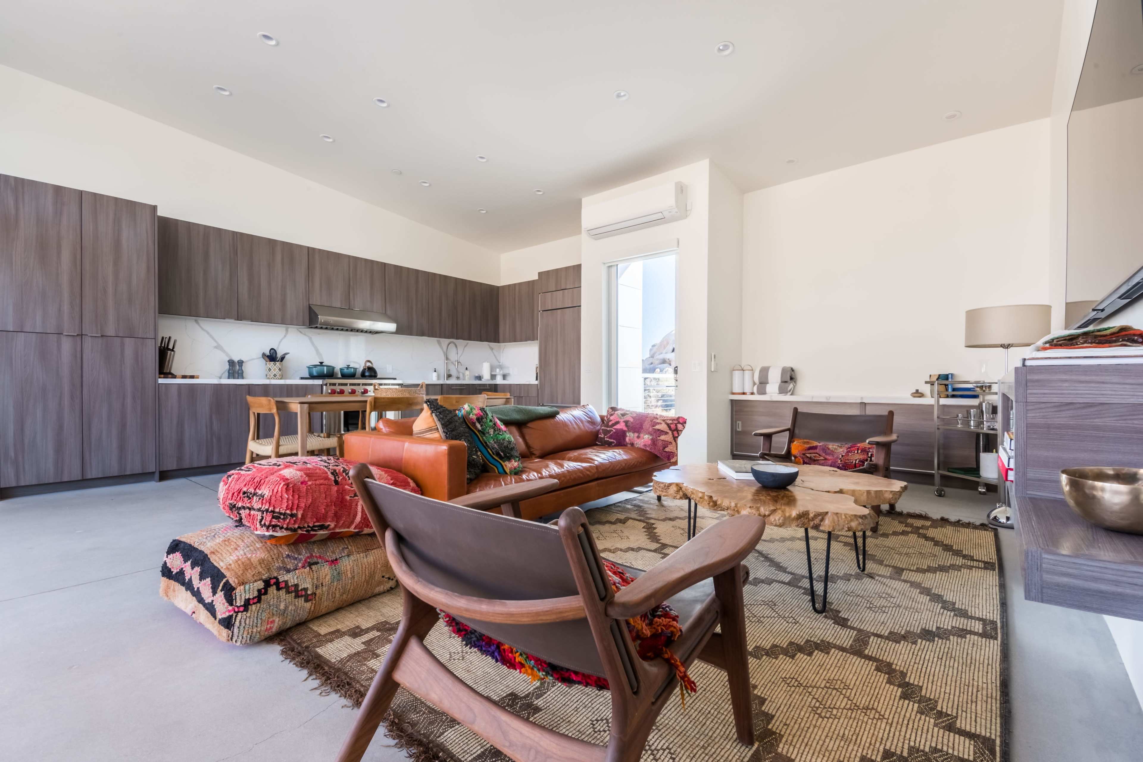 The image shows a modern living space with a kitchen area, a brown leather sofa, wooden furniture, and an area rug on the floor.