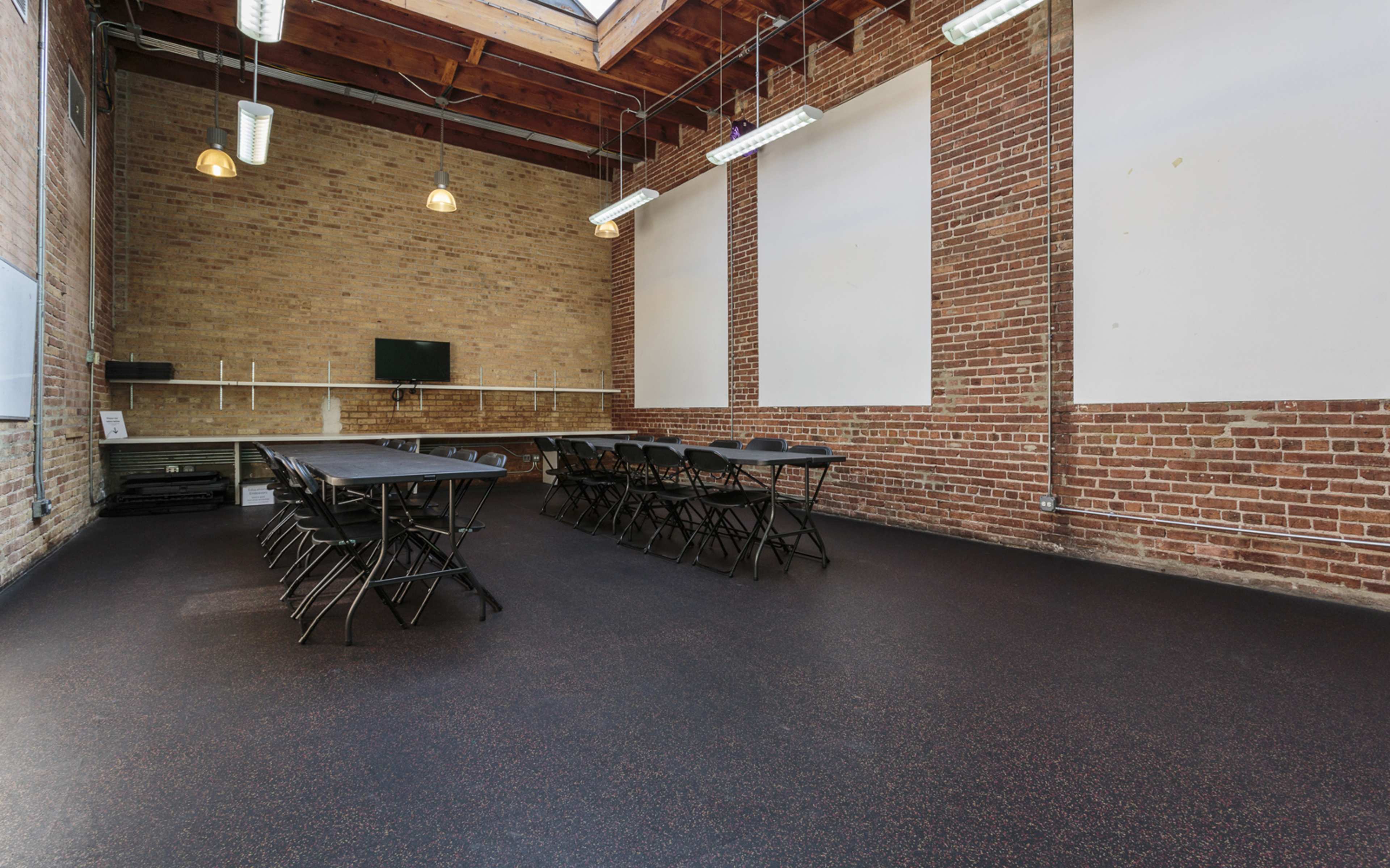 A spacious meeting room with a long black table surrounded by folding chairs, brick walls, and large windows overhead.